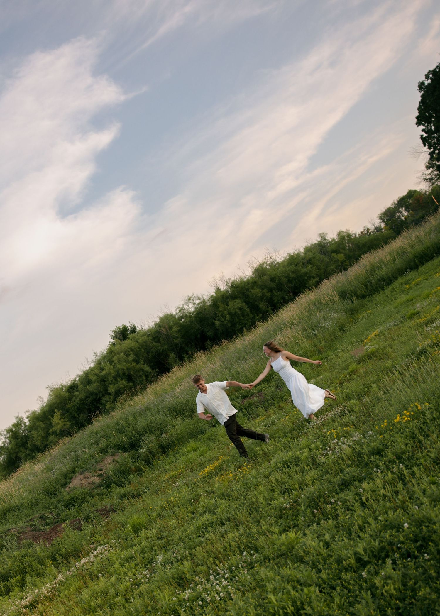 A couple in white attire holds hands while running through a lush green meadow under a cloudy sky.