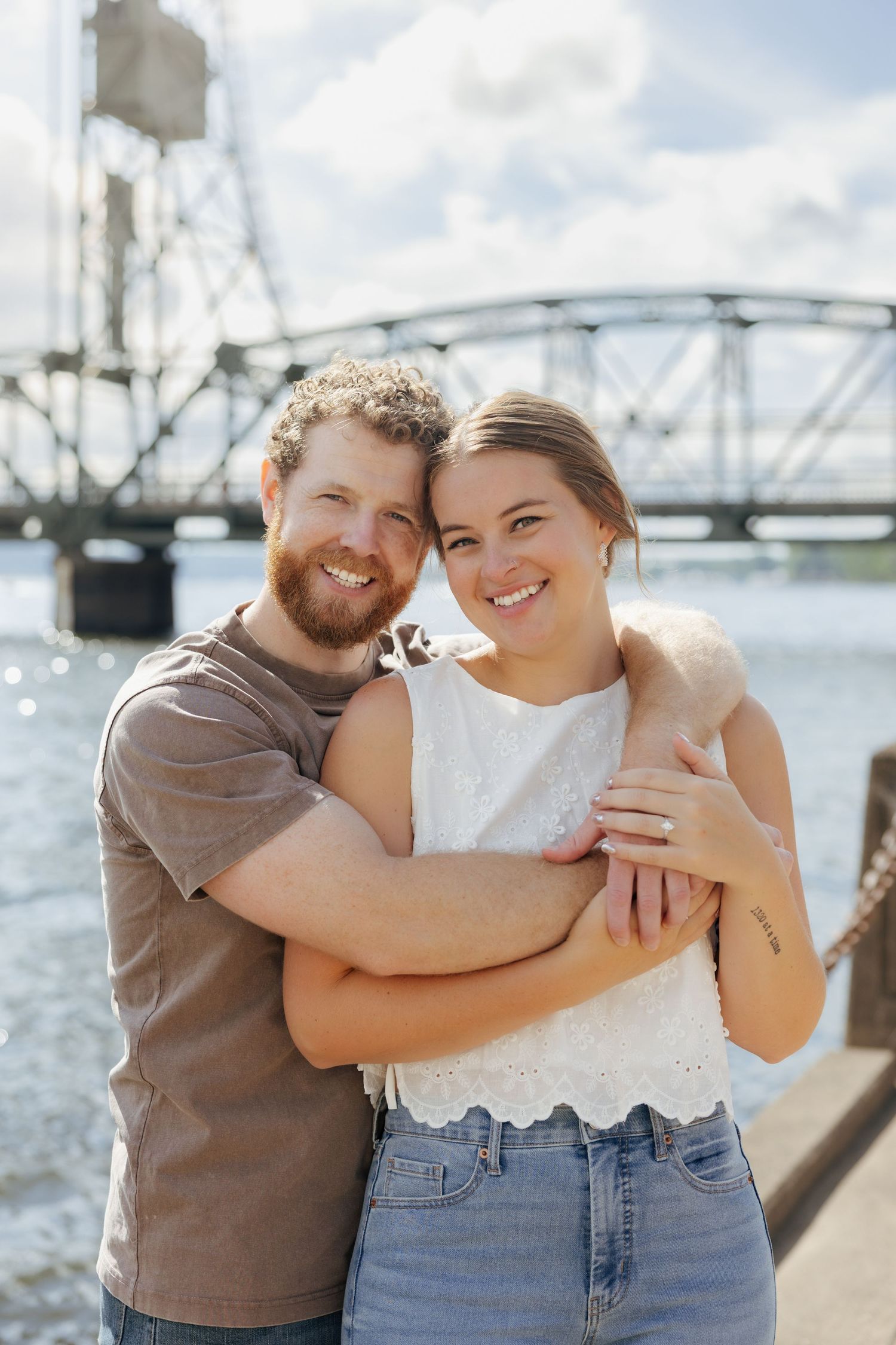 Happy couple embracing and smiling on a sunny waterfront dock with a steel lift bridge visible behind them.