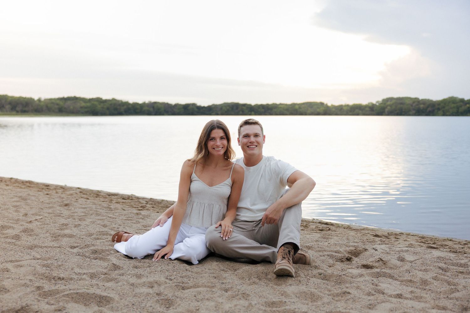 Smiling couple sitting together on sandy beach shore by calm lake at sunset in white and beige outfits.
