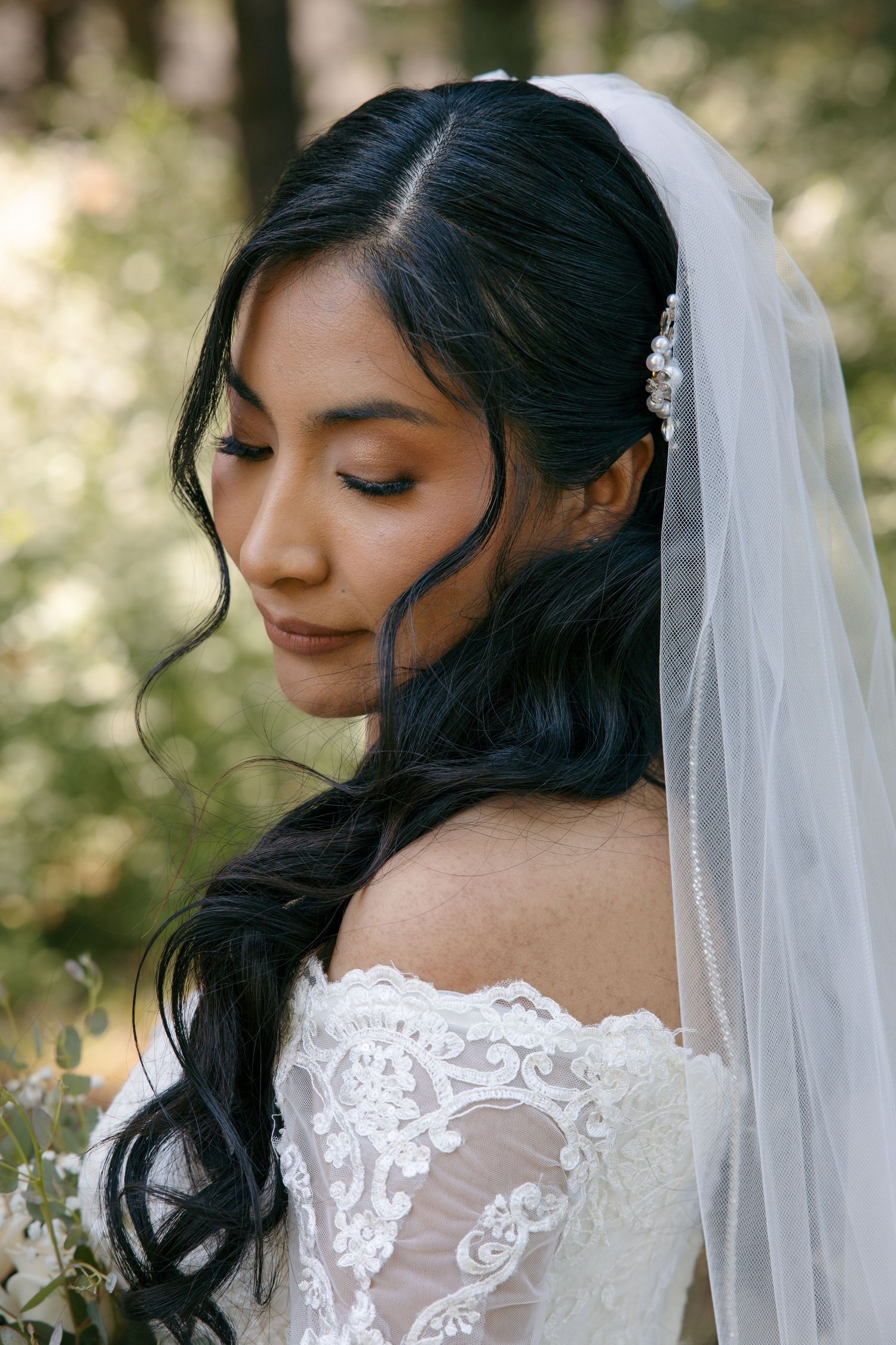 A bride in an off-shoulder lace gown and veil looks down gracefully among lush greenery.