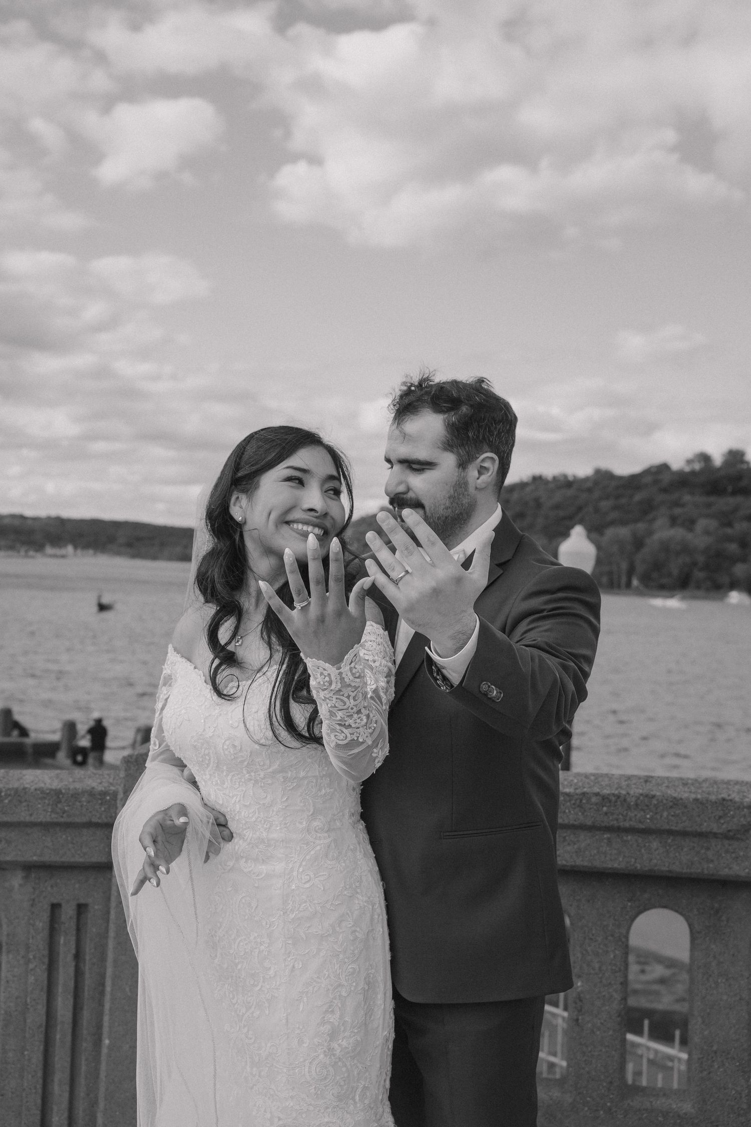 Black and white photo of newlyweds showing off their wedding rings to the camera with a lake in the background.