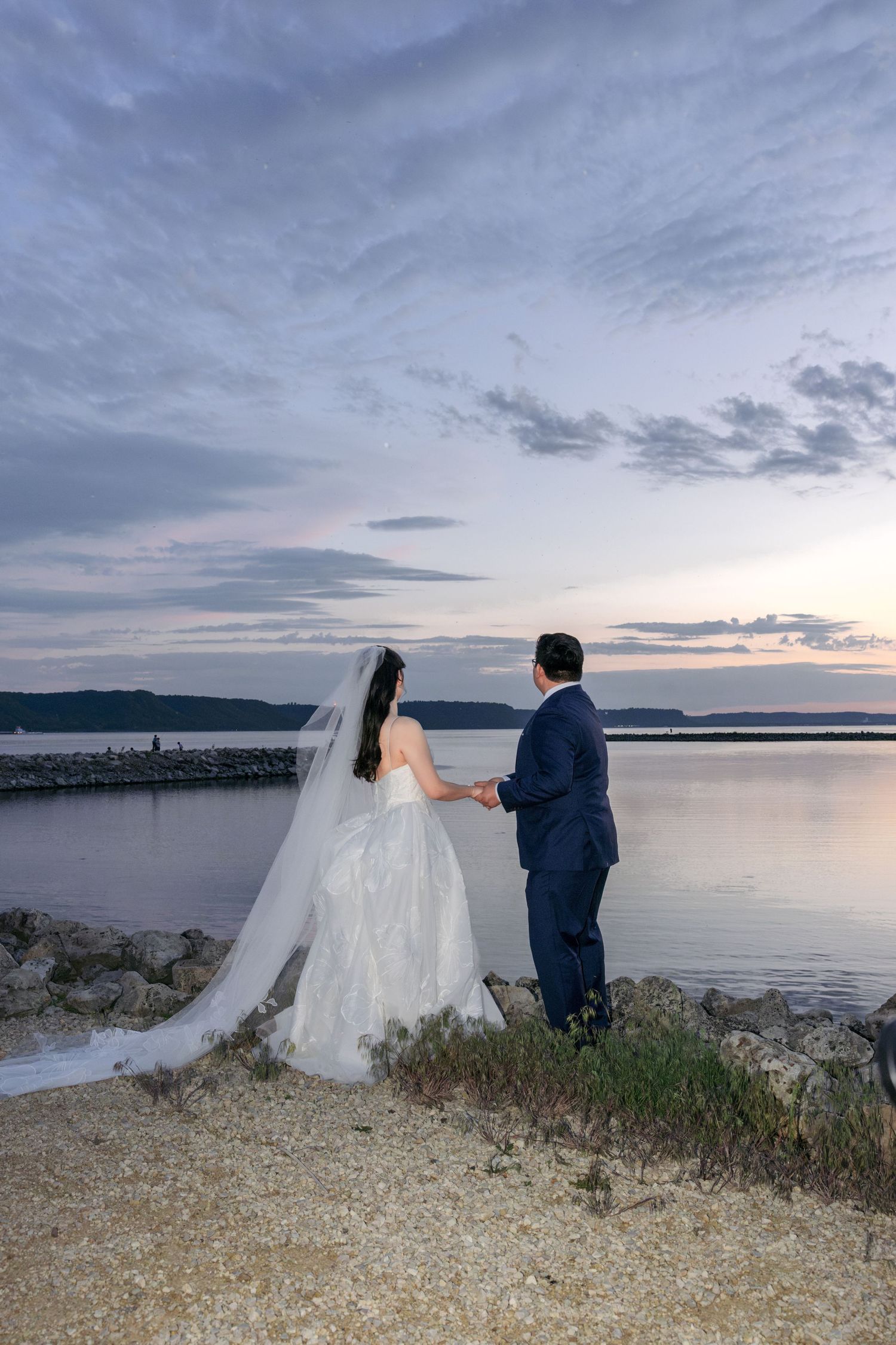 Bride and groom hold hands watching sunset over calm water at romantic outdoor waterfront wedding.
