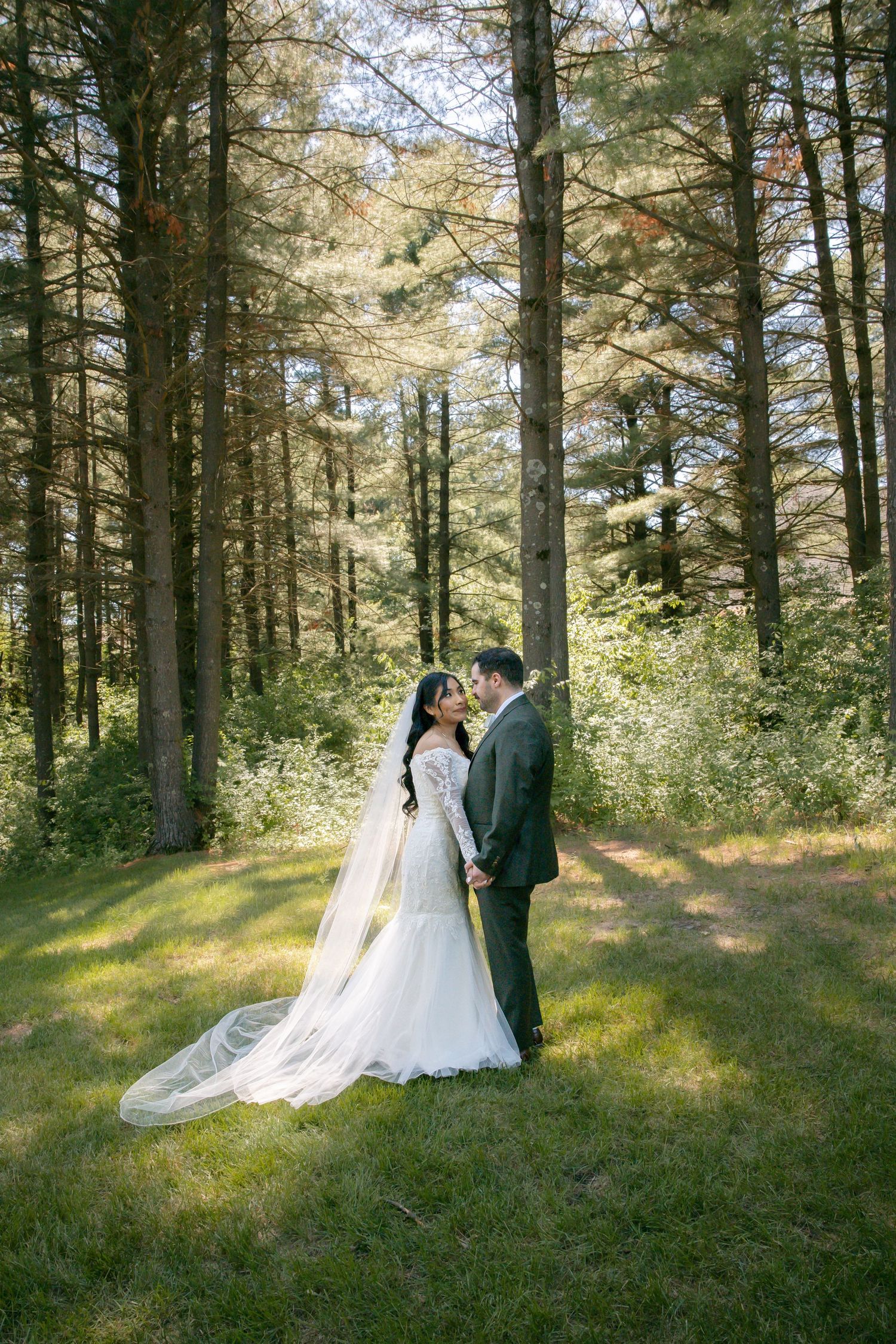 Bride and groom share a romantic moment in a sunlit forest clearing, bride wearing a lace gown with flowing veil.