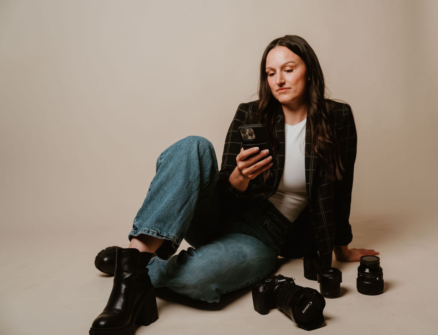 Woman sitting on floor in black blazer and jeans, looking at smartphone with camera nearby on neutral background.