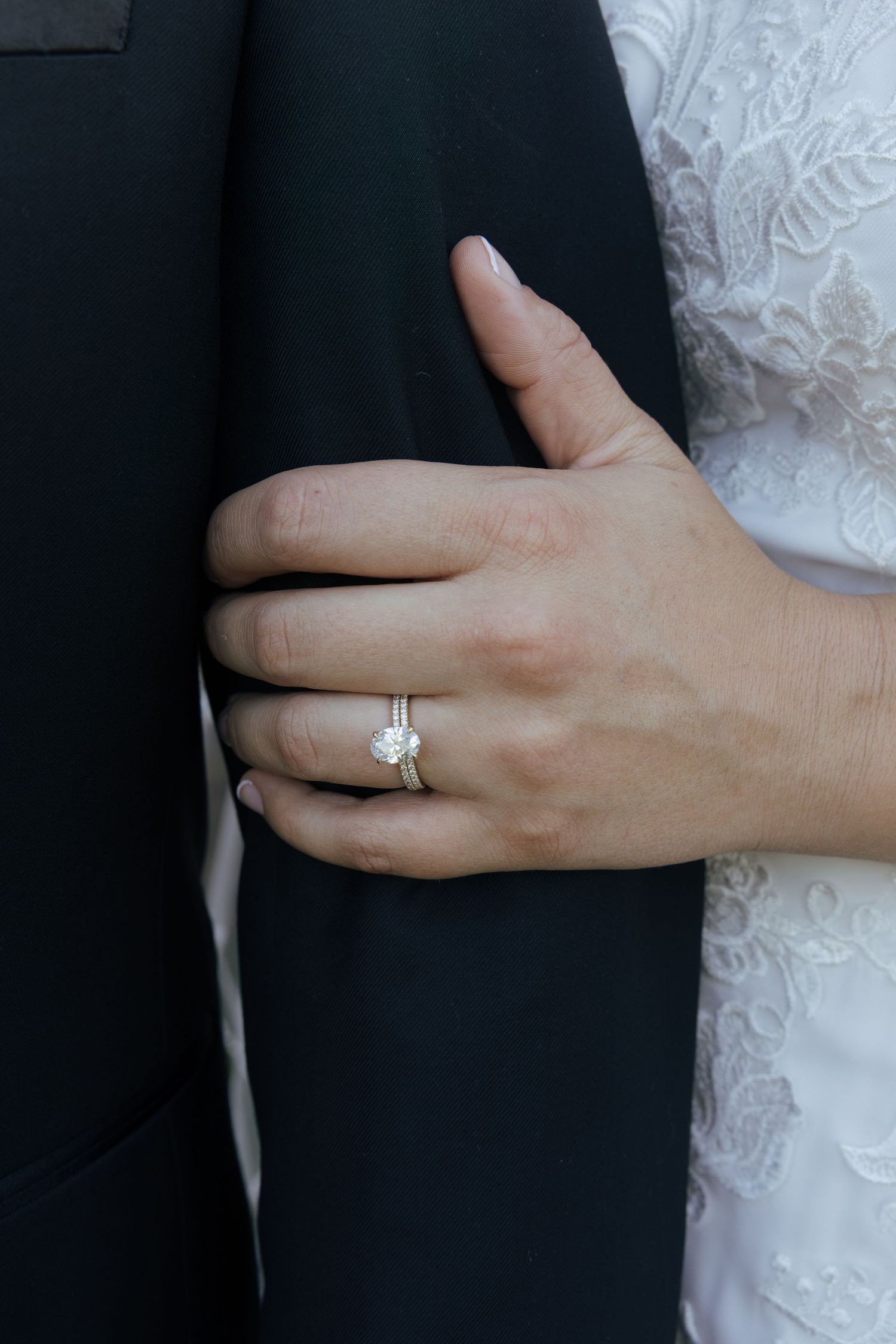 Close-up of a bride's hand with a solitaire diamond engagement ring resting on a groom's black suit jacket.