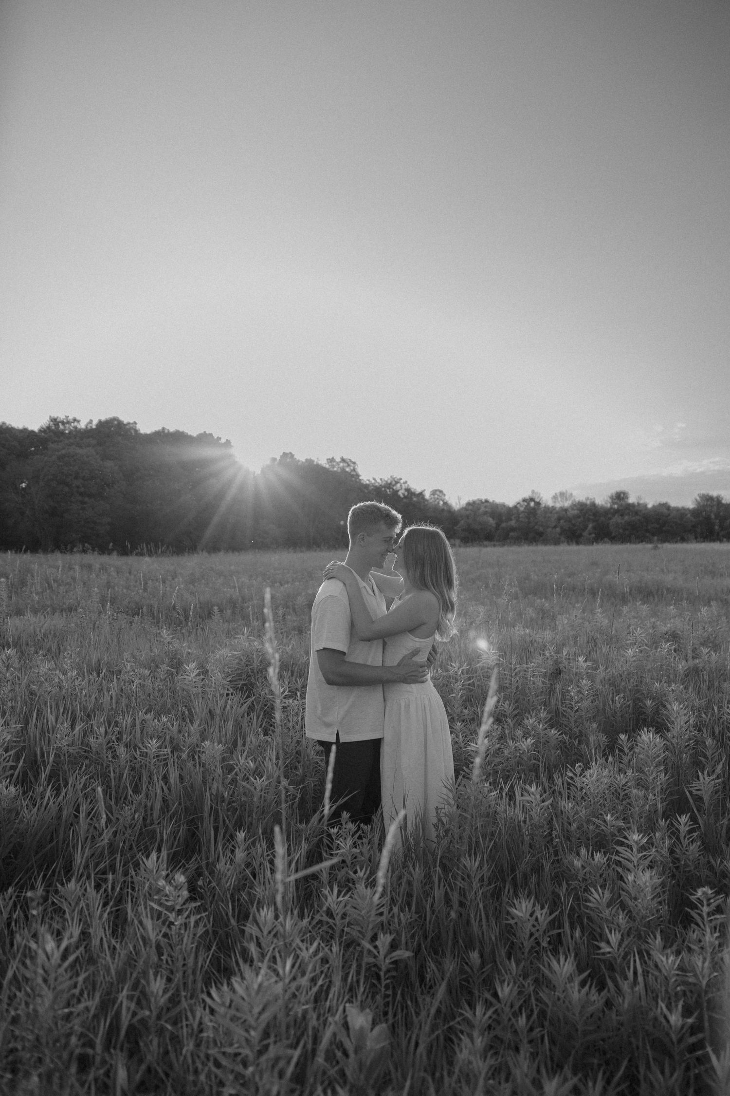 Black and white photo of a couple embracing in a lush field at sunset with sun rays behind them.
