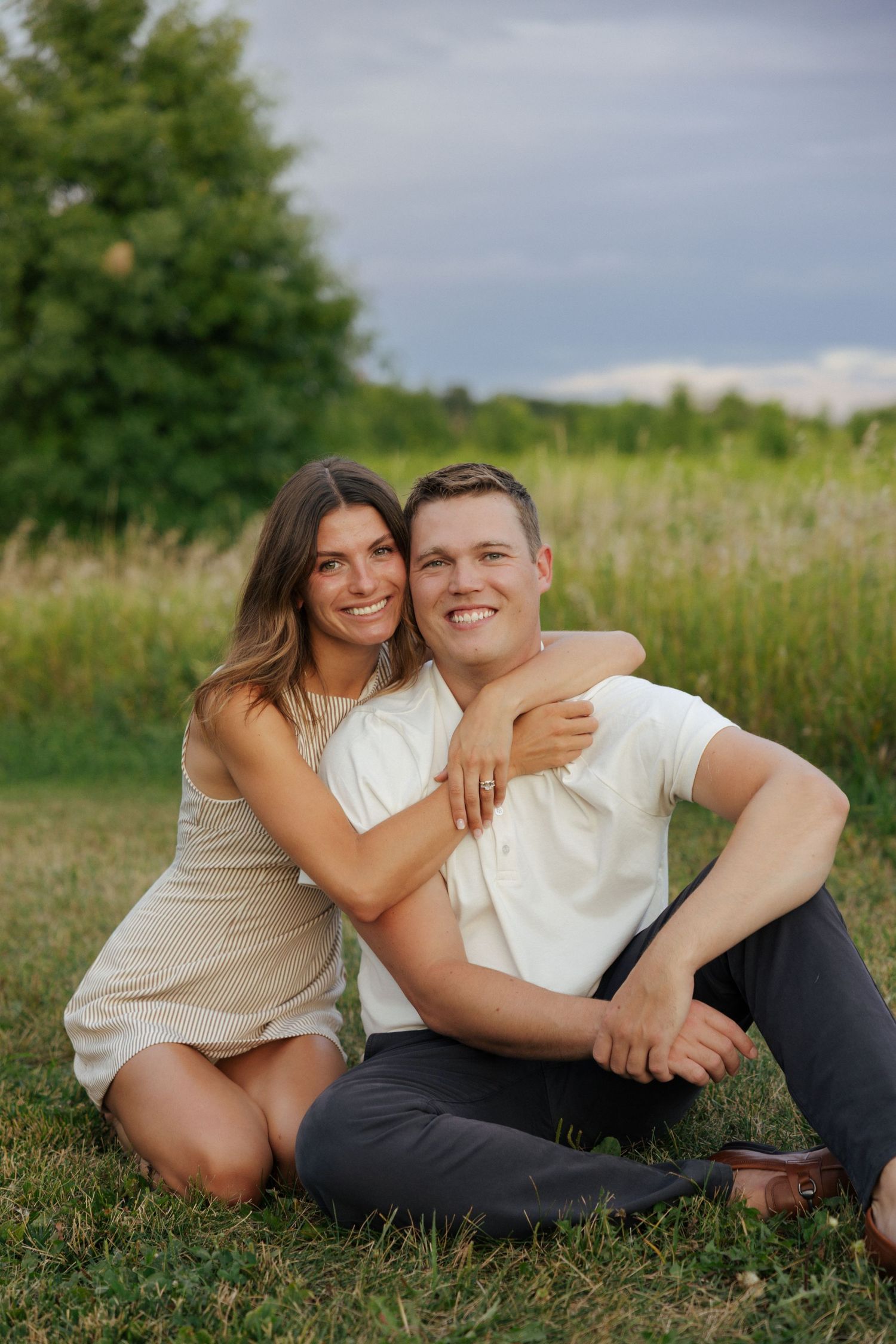 A smiling couple sits together on green grass with trees in the background during golden hour.