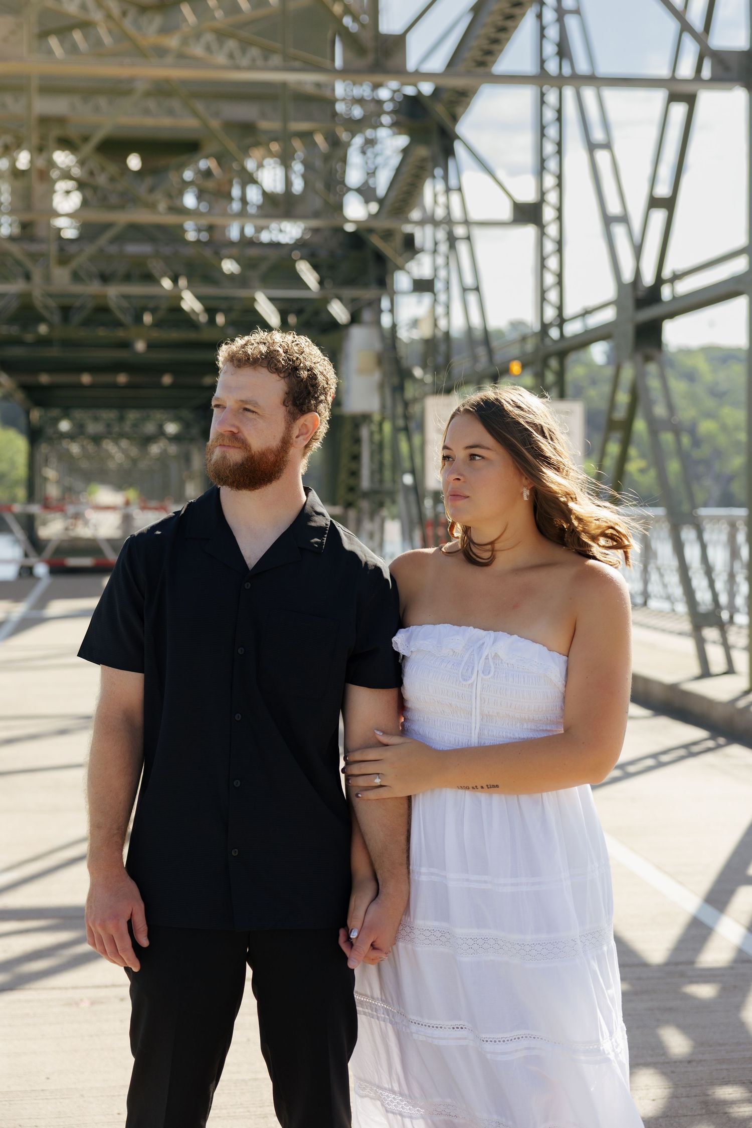 A bearded man in black and a woman in a white strapless dress pose together on a bridge on a sunny day.