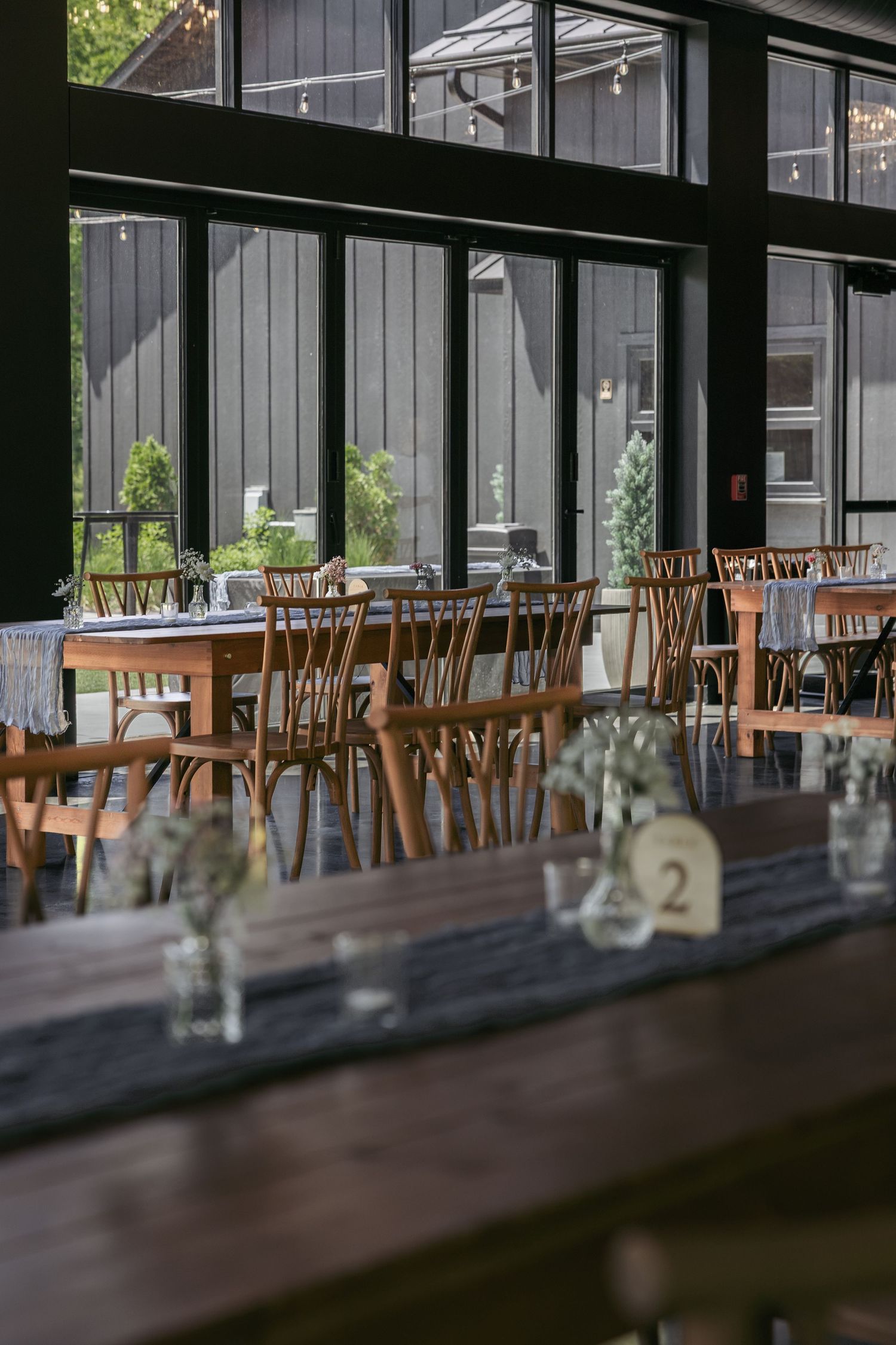 Rustic wedding reception venue with wooden tables, cross-back chairs, and floral centerpieces in a barn-style setting.