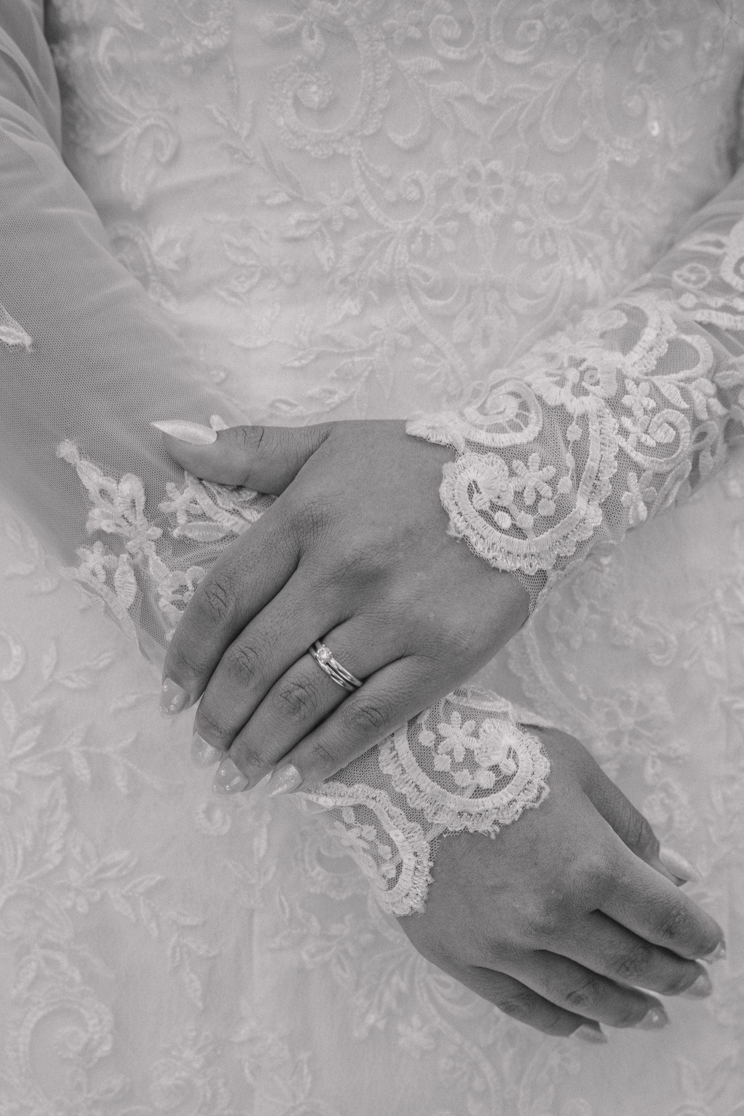 Black and white detail shot of brides hands adorned with wedding ring resting on intricate lace wedding gown.