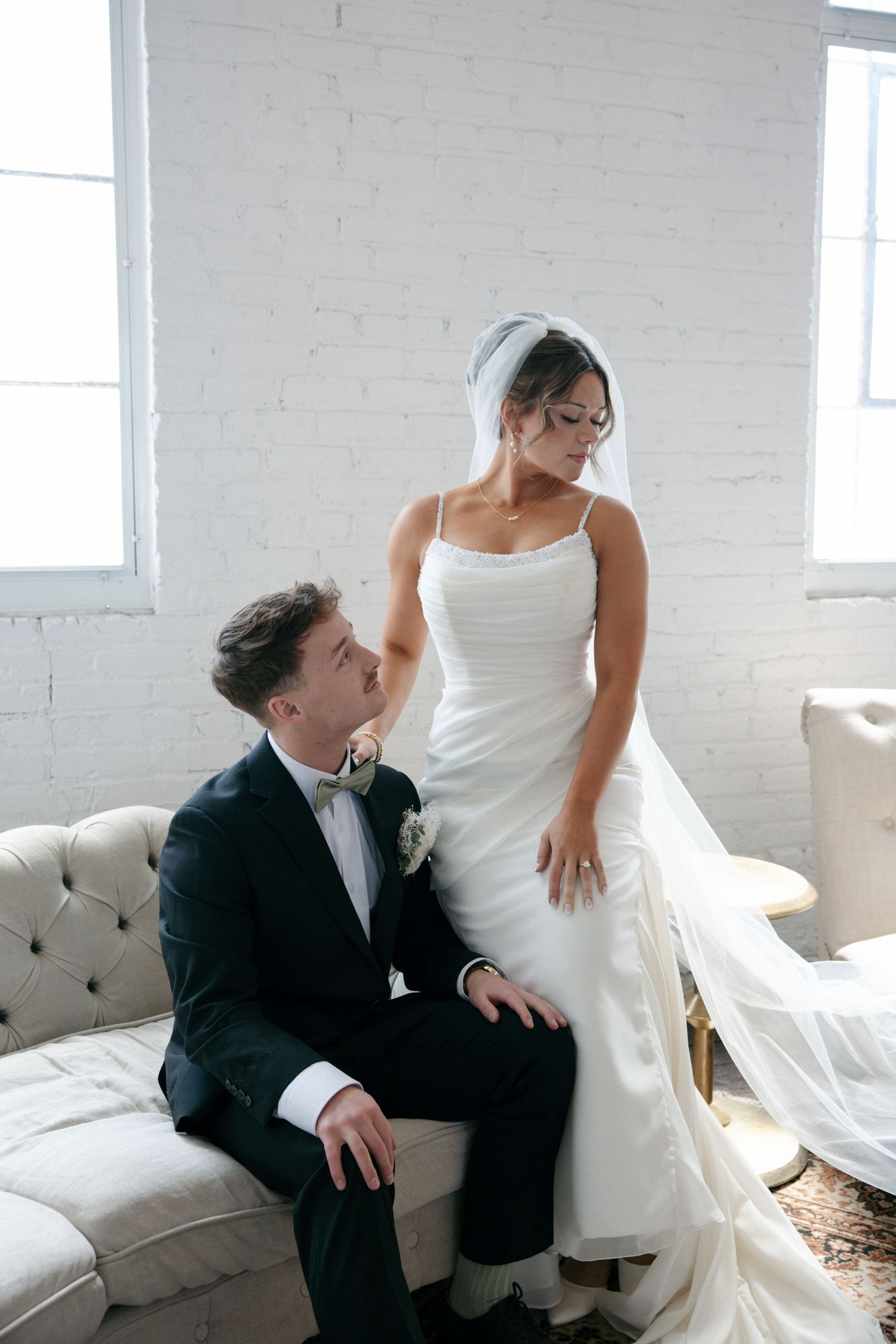 Bride and groom share an intimate moment on a tufted sofa, with the bride in a sleek white gown and veil.