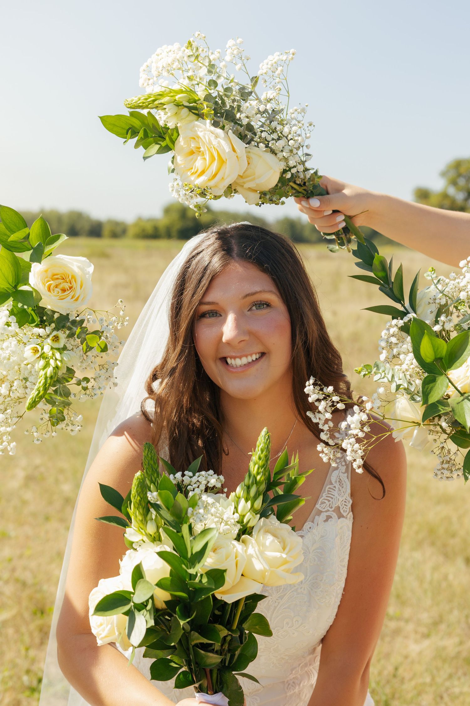 Smiling bride in lace gown and veil surrounded by white rose and greenery bouquets in sunny field.