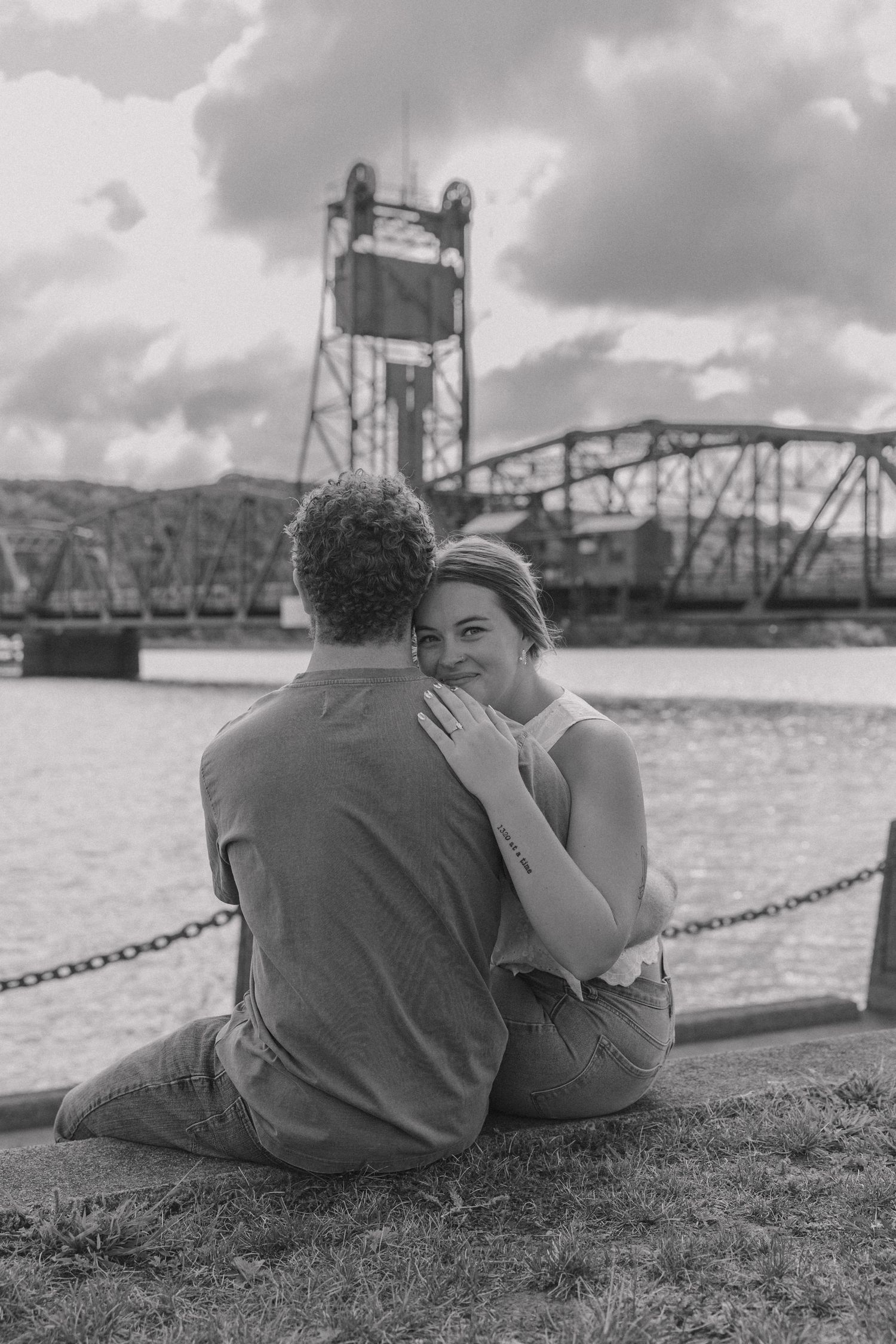 Black and white photo of a couple sitting by a river, woman smiling over mans shoulder with a lift bridge behind them.