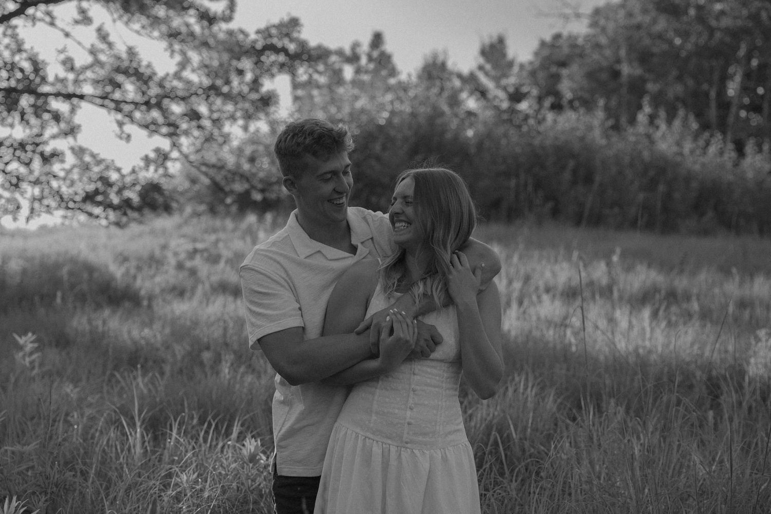 Black and white photo of a couple embracing and smiling in a field with trees in the background.
