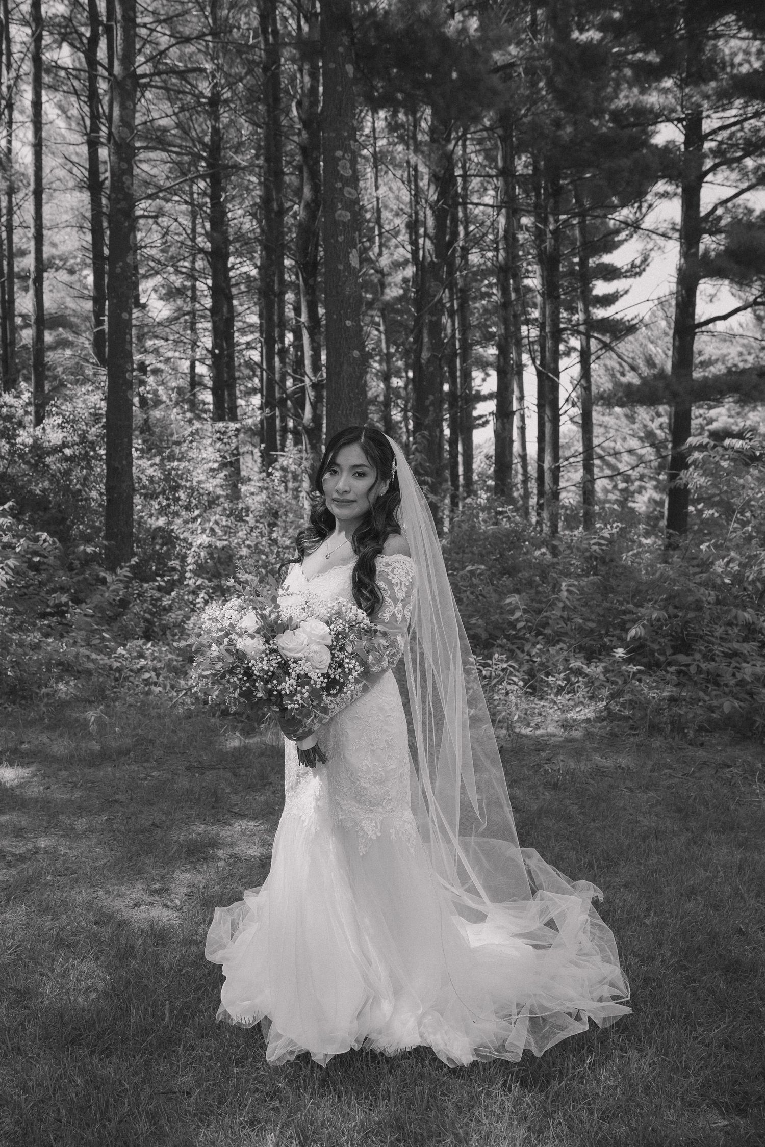 Bride in white lace gown and veil holds bouquet in black and white forest portrait.