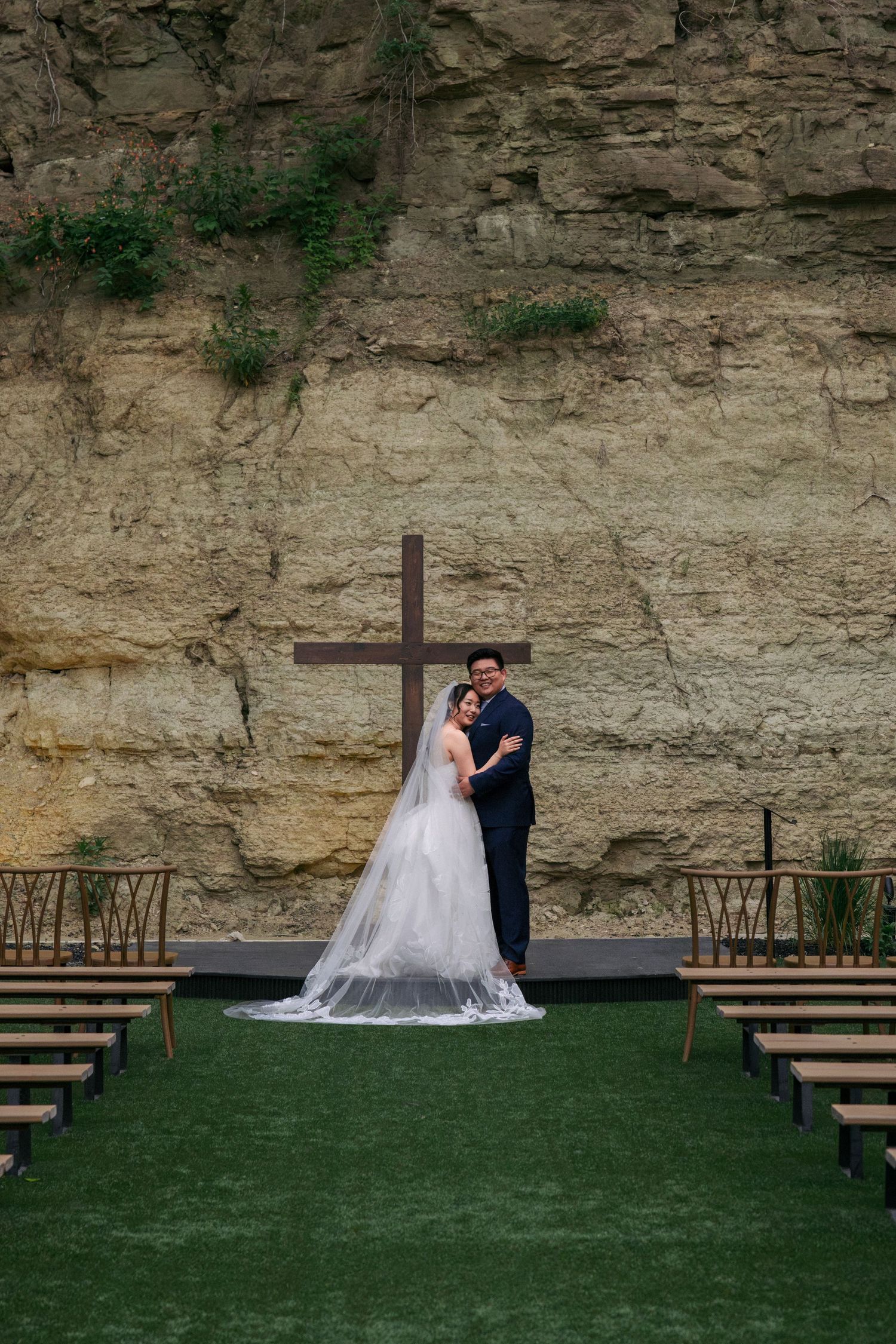 Bride and groom embrace before a wooden cross set against a stunning natural limestone cliff backdrop.