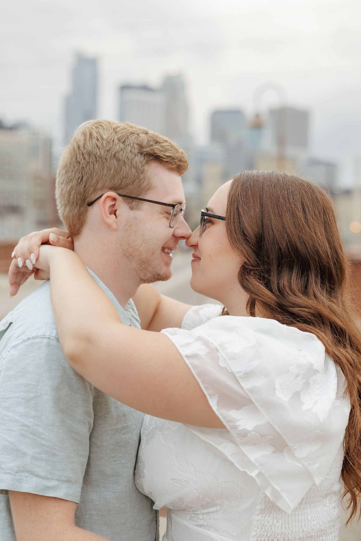 Couple sharing a romantic nose-to-nose moment with city skyline backdrop, woman in white top embracing man in gray tee.