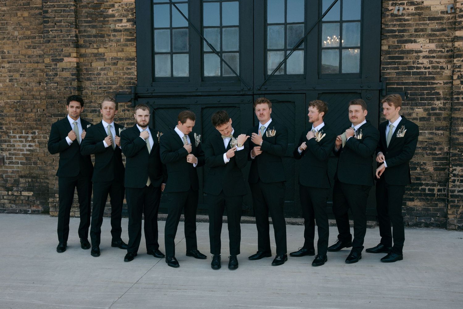 Nine groomsmen in matching black suits posing and adjusting cufflinks outside a brick building.