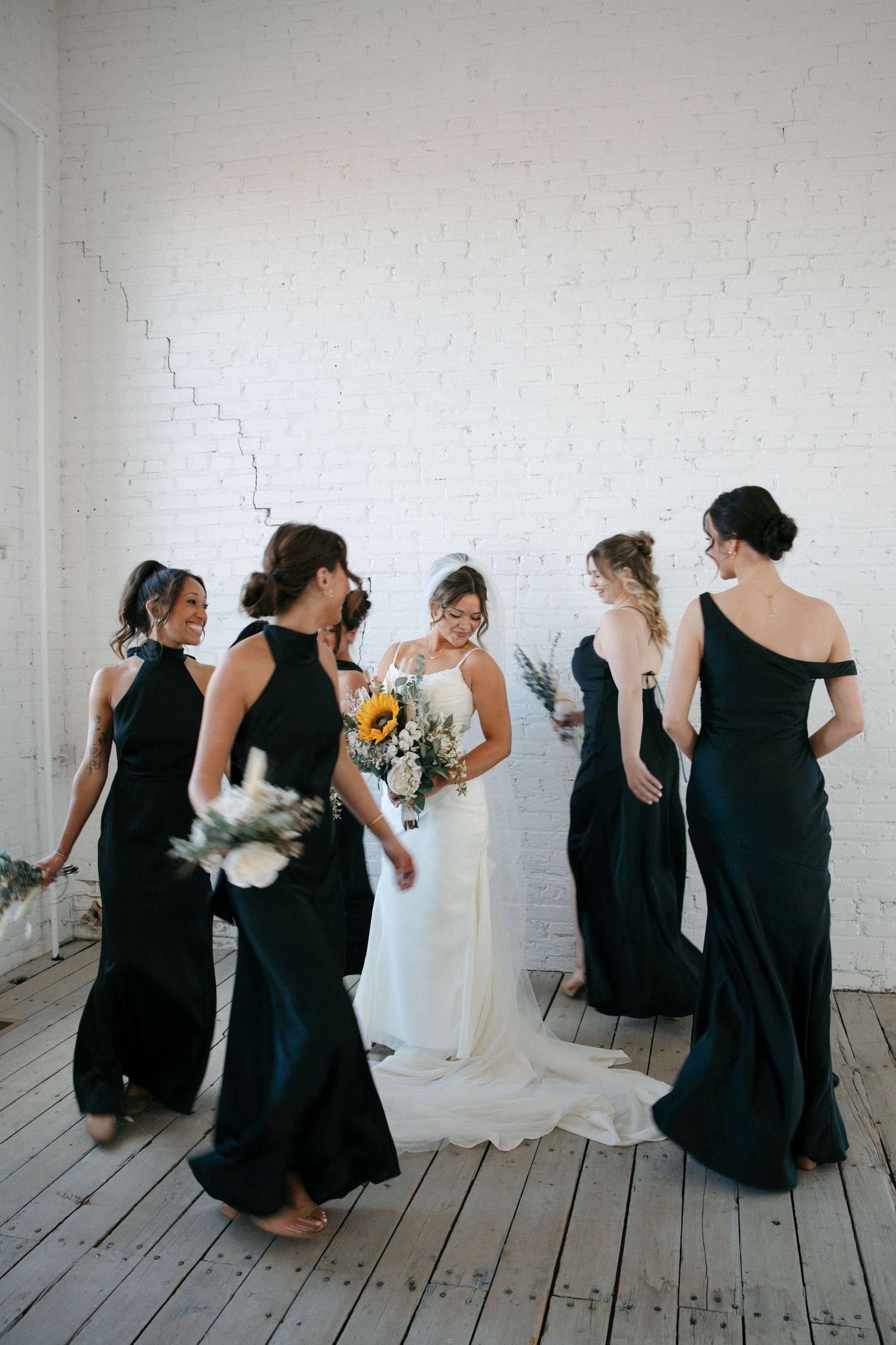 Bride in white dress surrounded by bridesmaids in black gowns holding bouquets against white brick wall.