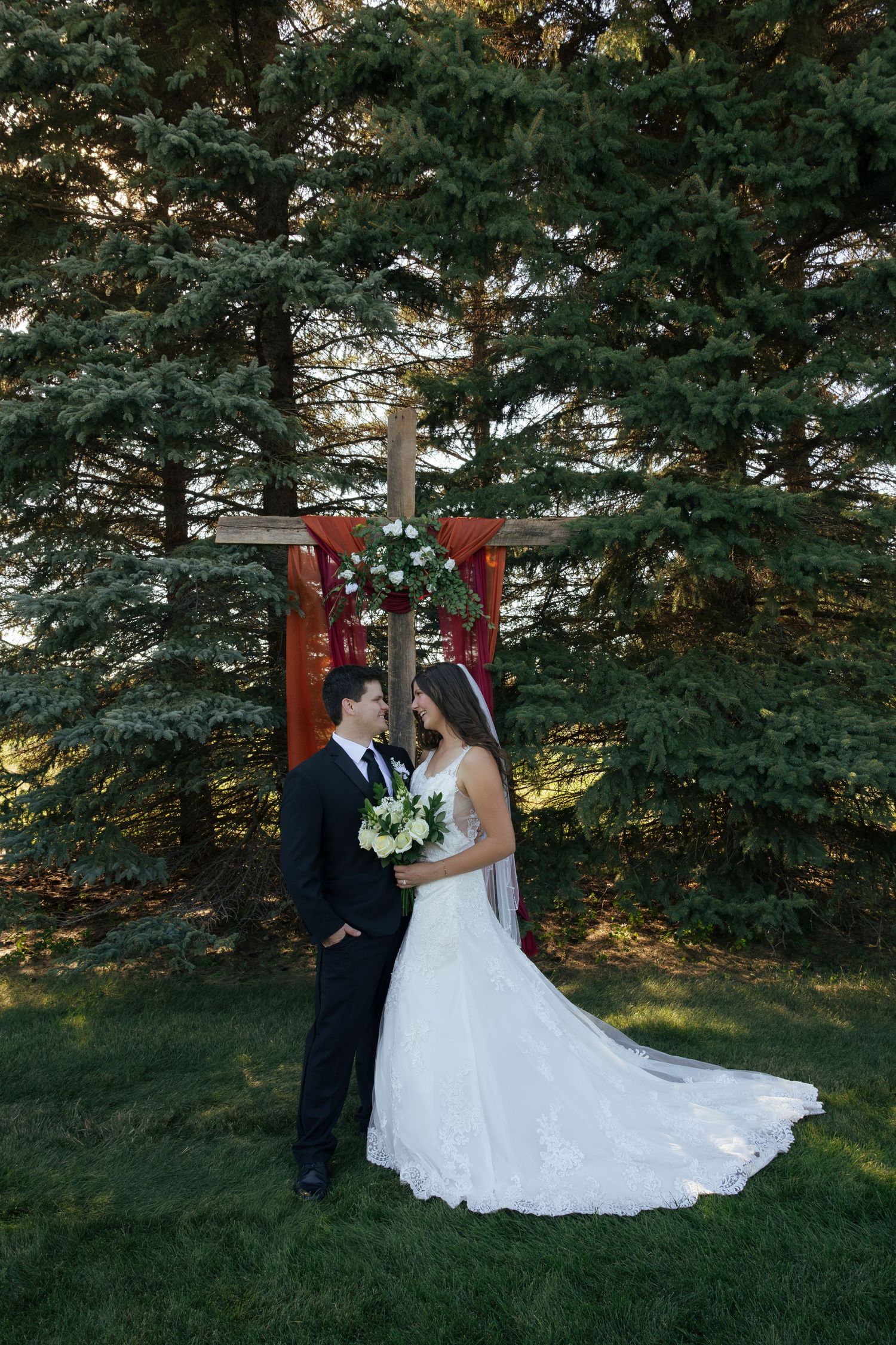 Bride and groom sharing romantic moment before wooden cross altar surrounded by pine trees.