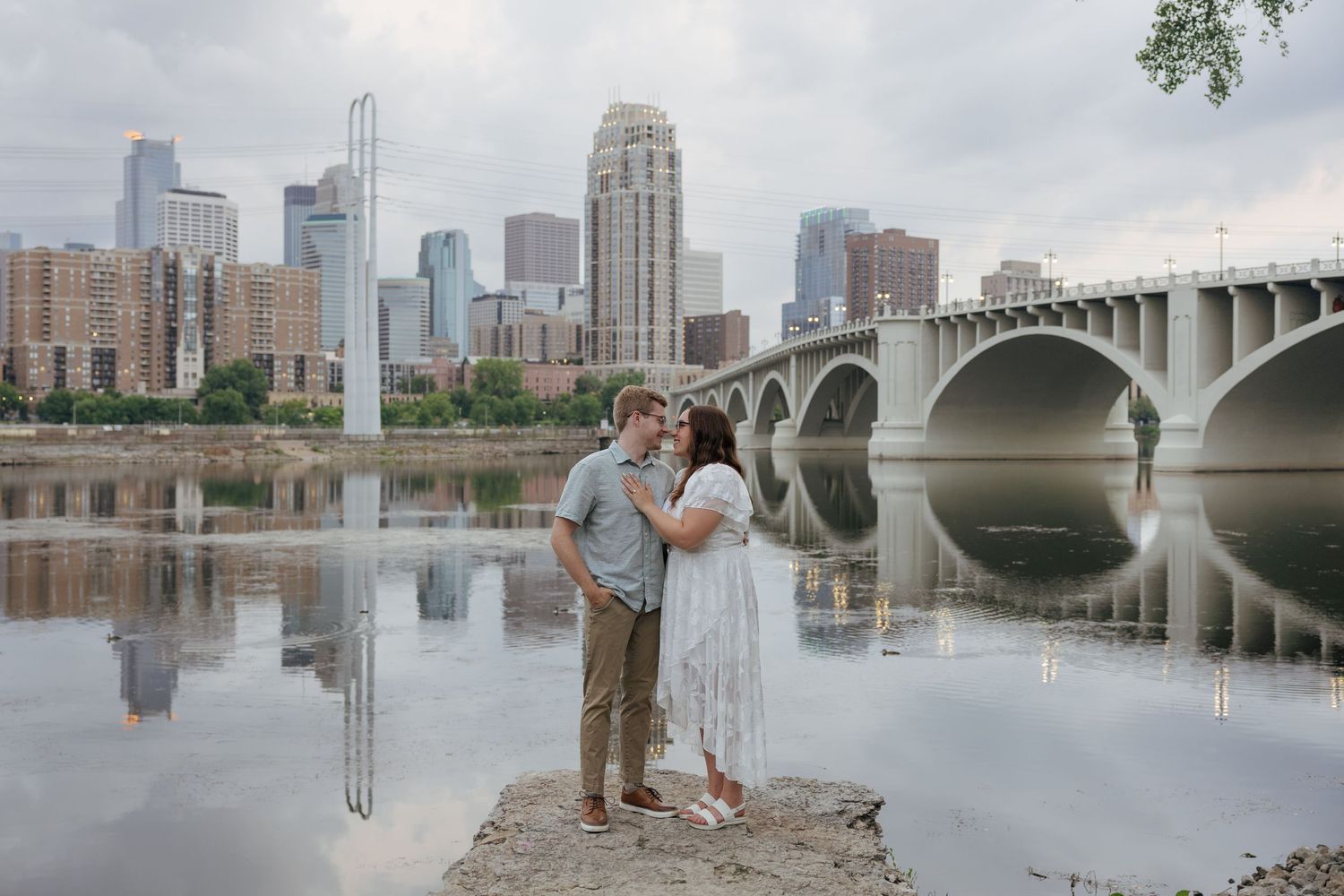 Couple sharing a romantic moment by the Mississippi River with Minneapolis skyline and bridge reflected in water.