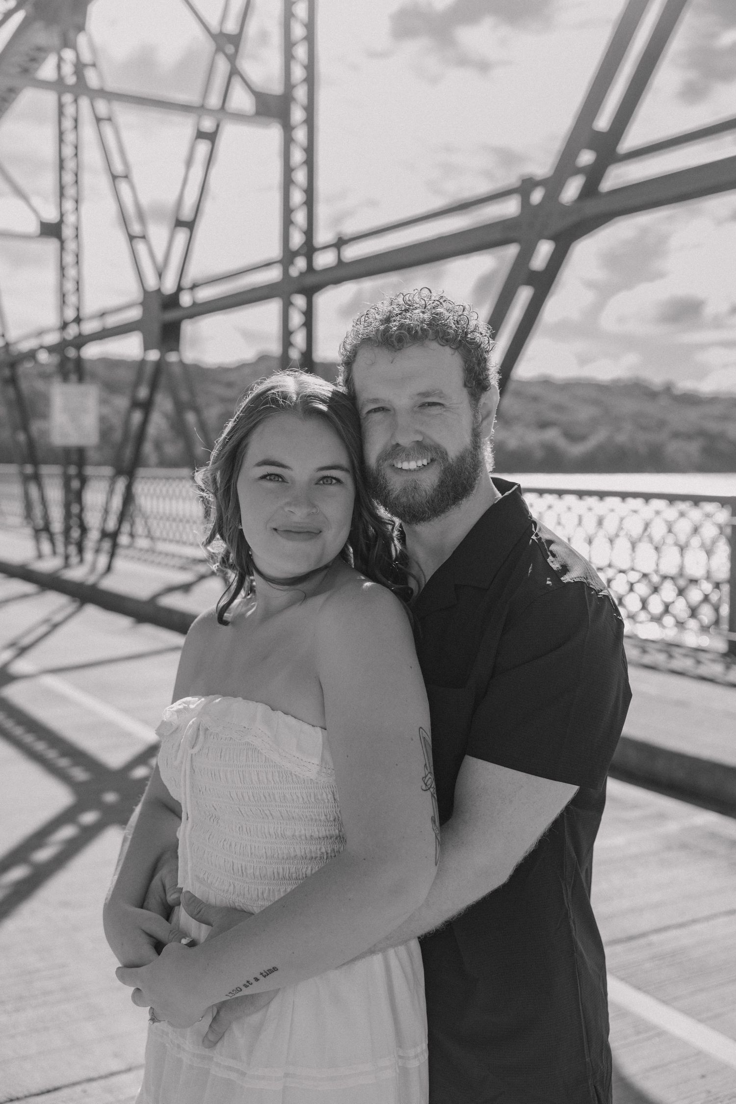 Black and white photo of a smiling couple embracing on a bridge, man with beard in black shirt, woman in white dress.