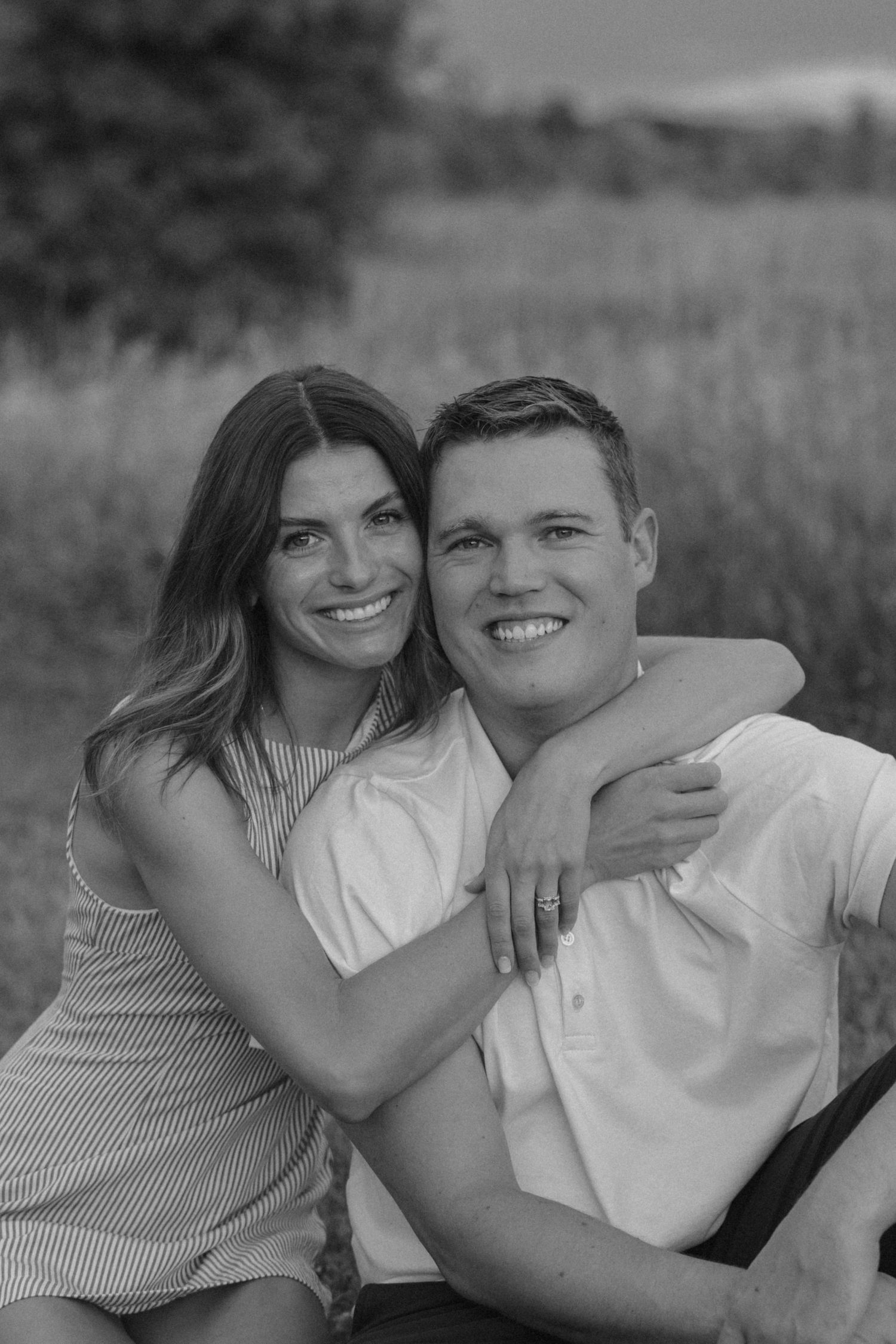 Black and white photo of a smiling couple embracing outdoors in a field, woman in striped dress hugging man in white shirt.