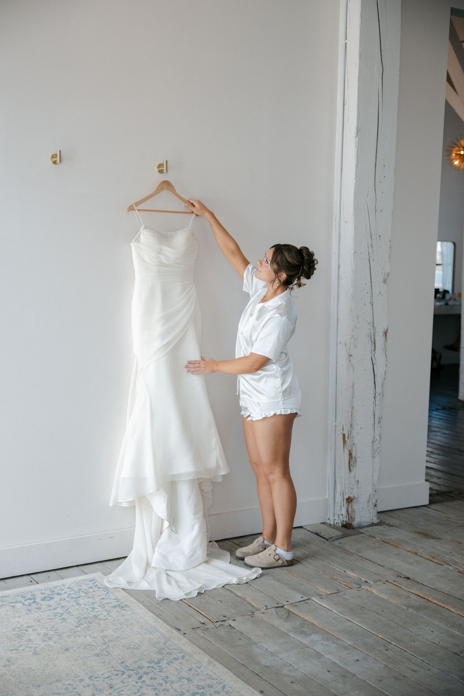Bride in white shorts set admiring wedding gown hanging on wall hook before ceremony.