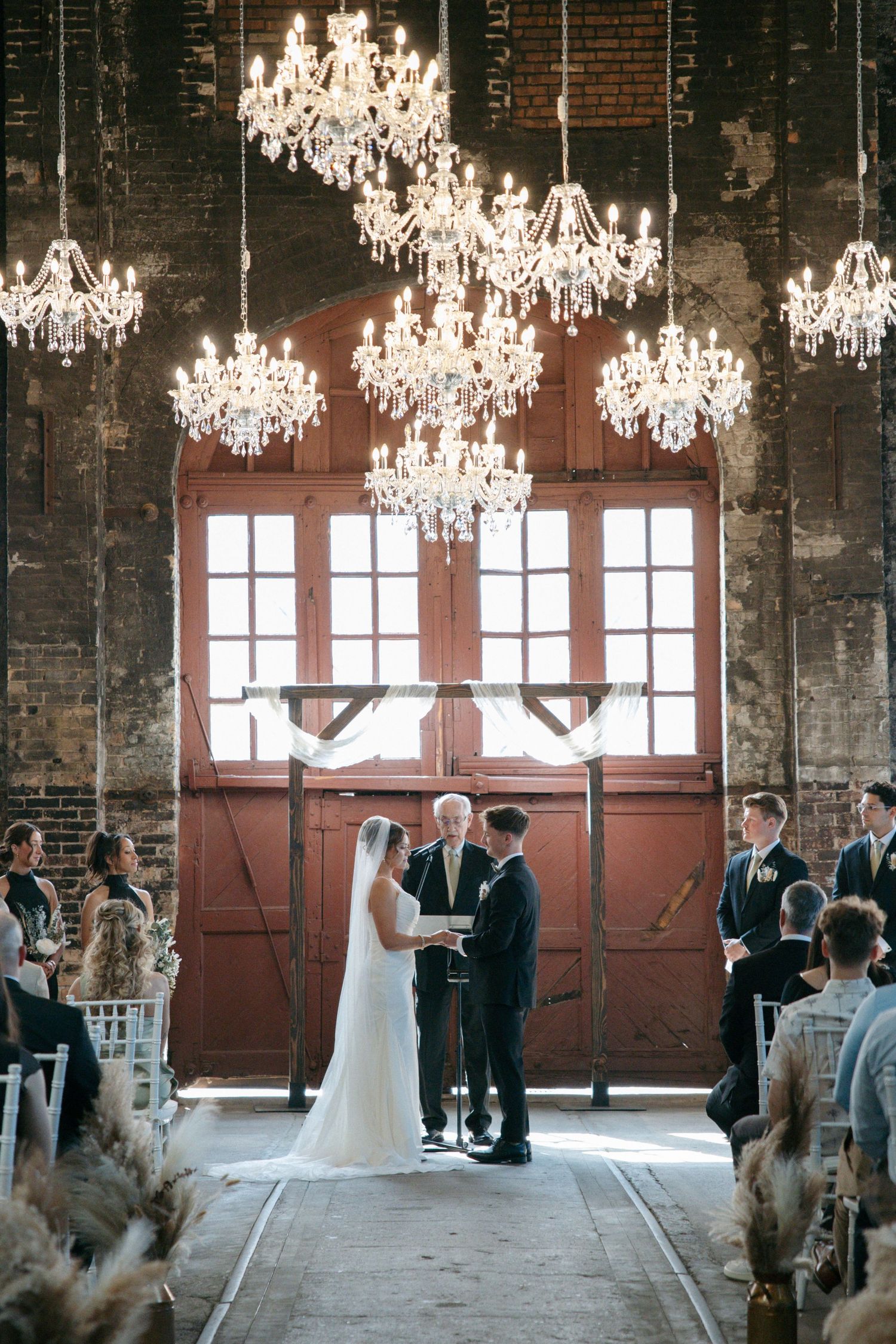 Bride and groom exchange vows under crystal chandeliers in a rustic industrial wedding venue with red barn doors.