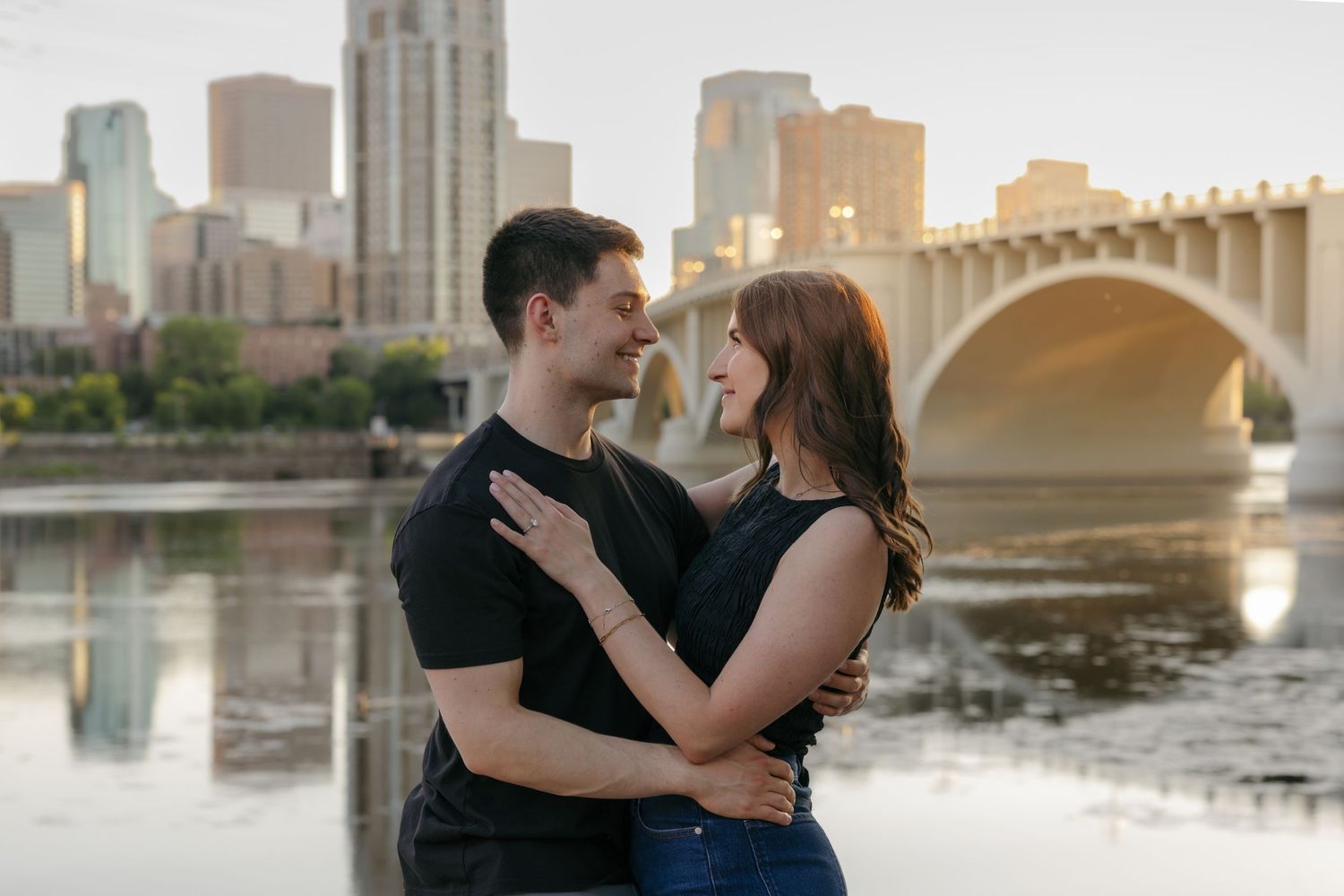 Couple embracing and smiling at each other with Minneapolis skyline and bridge reflected in river at golden hour.