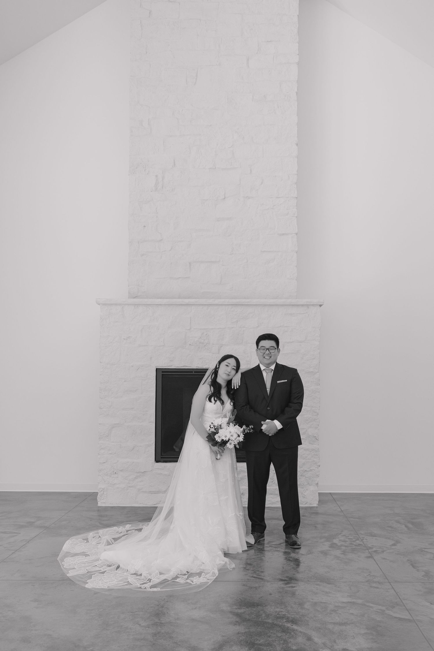 Black and white wedding photo of bride and groom standing before a white stone fireplace, bride holding bouquet.