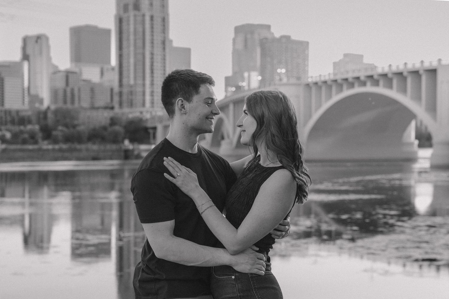 Black and white photo of a couple embracing and smiling at each other with a city skyline and bridge in the background.