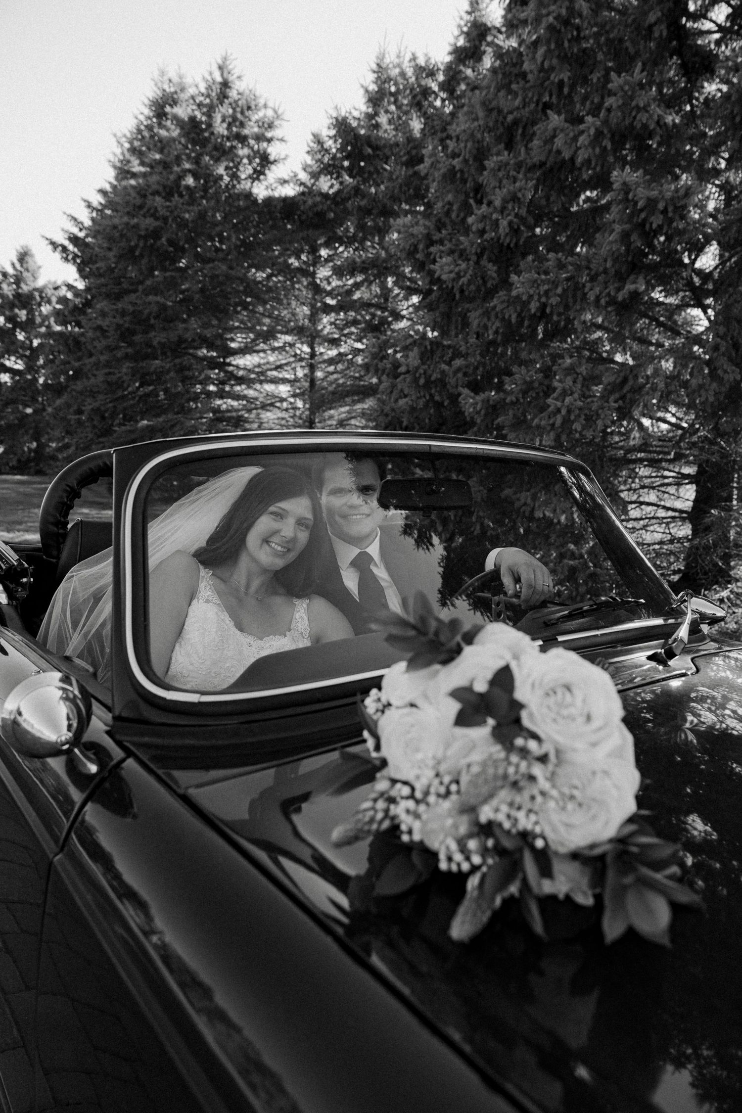 Black and white photo of bride and groom in vintage convertible with floral bouquet.
