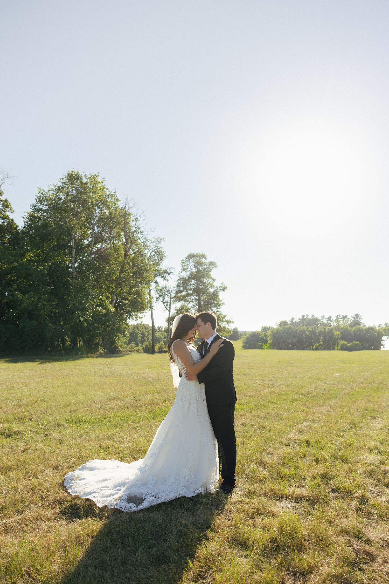 Bride and groom embrace in a sunlit green field, bride in white gown with train, groom in black suit.