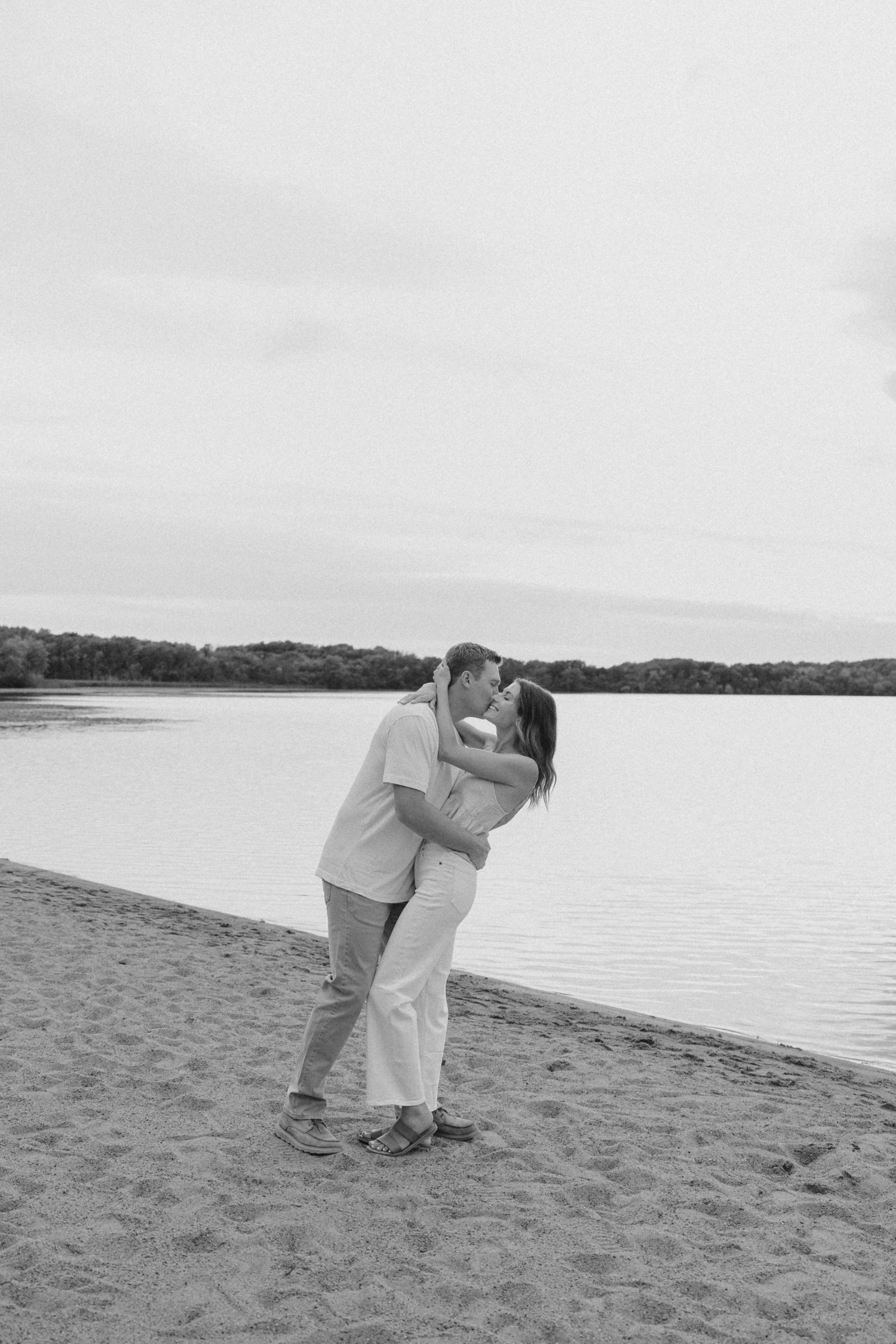 Black and white photo of a couple kissing on a sandy beach shore with a calm lake and trees in the background.