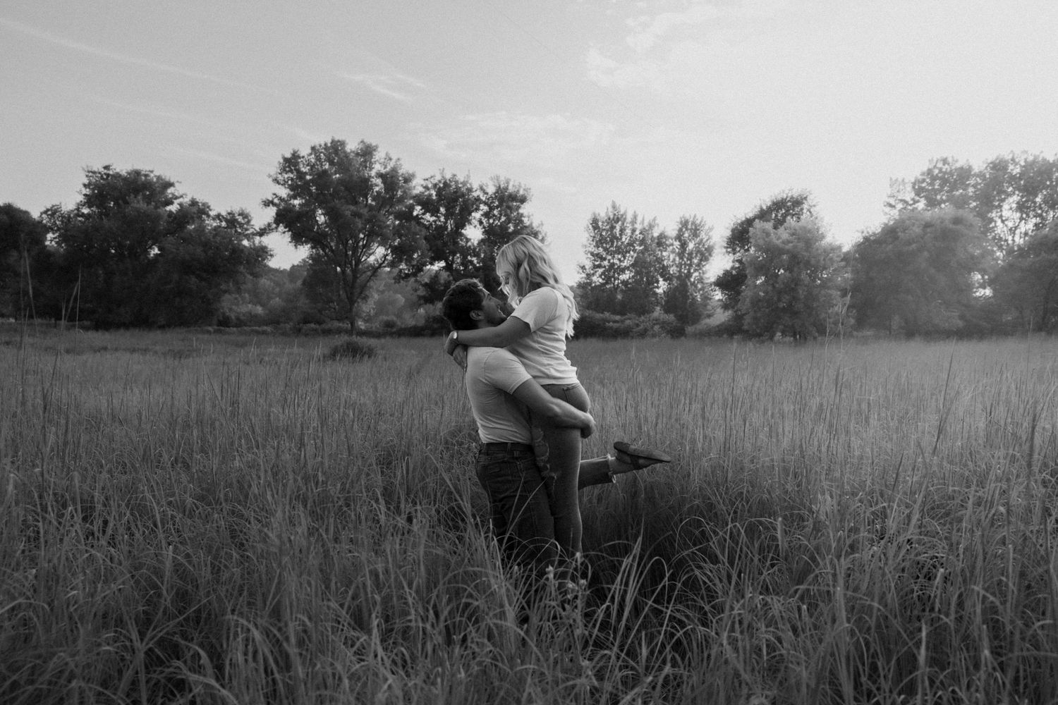 Black and white photo of a couple embracing in a tall grass field surrounded by trees.