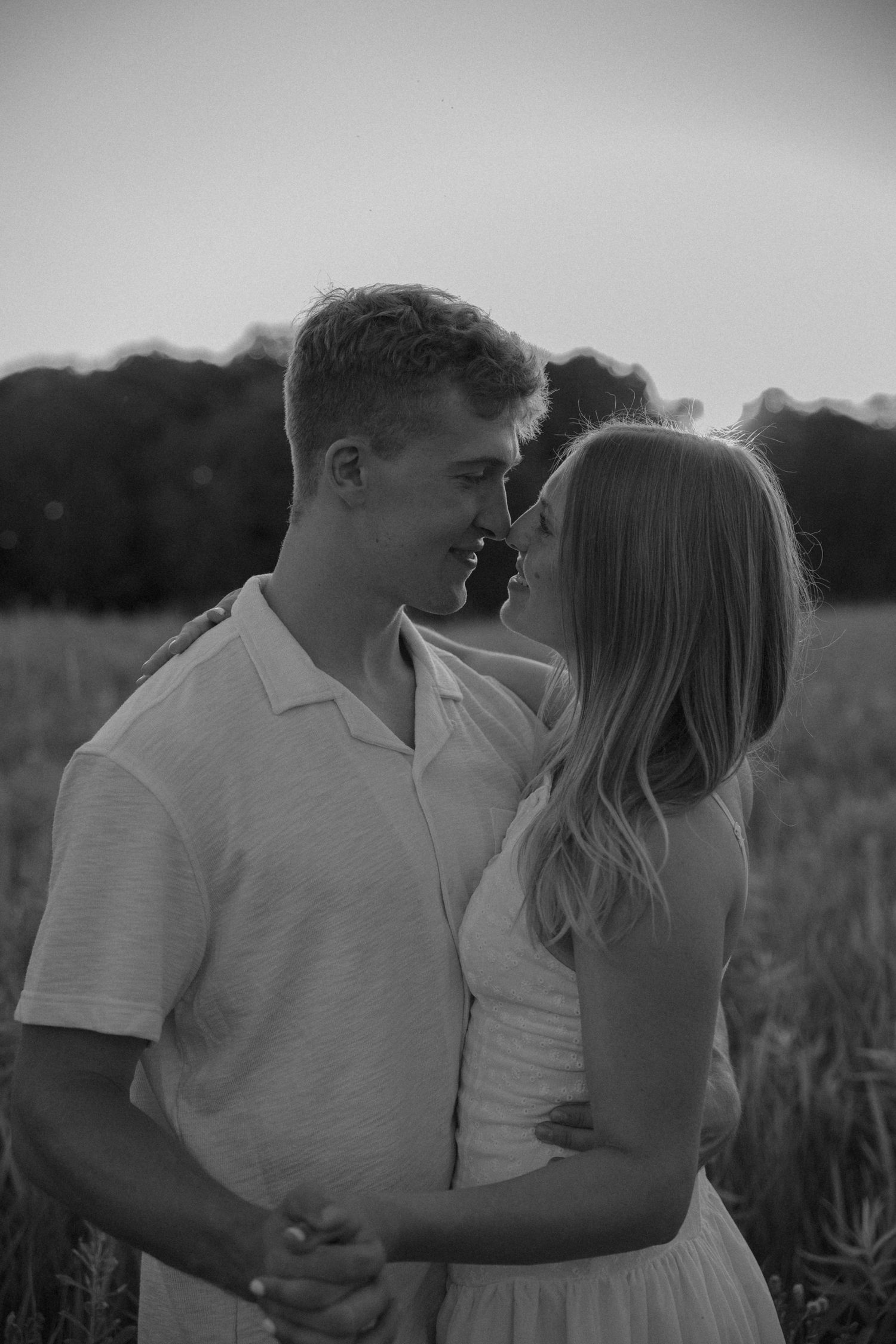 Black and white photo of a young couple sharing a romantic moment in a field at dusk, smiling and holding hands.