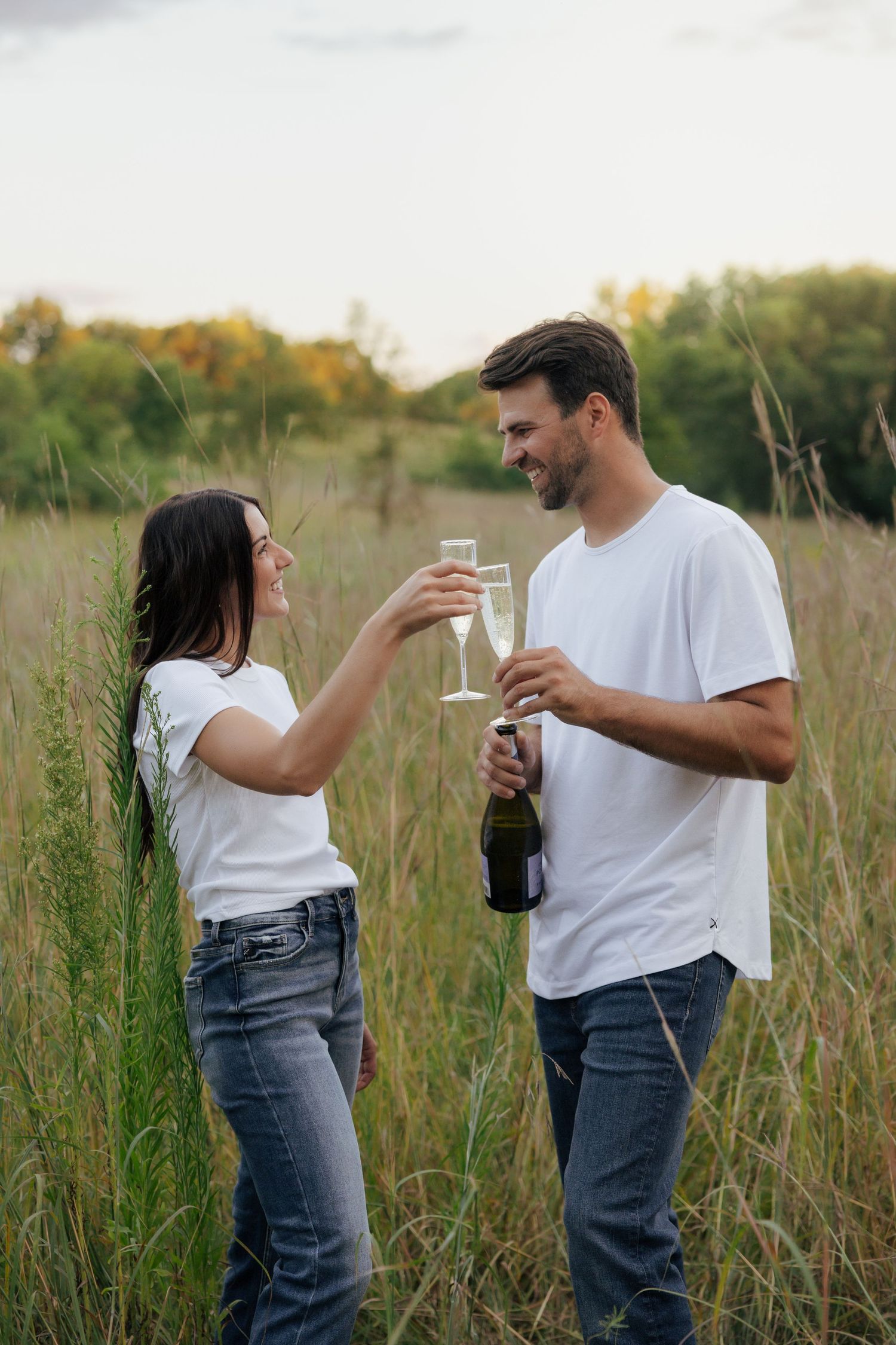 Couple in white t-shirts toasting with champagne glasses in a tall grass field outdoors.