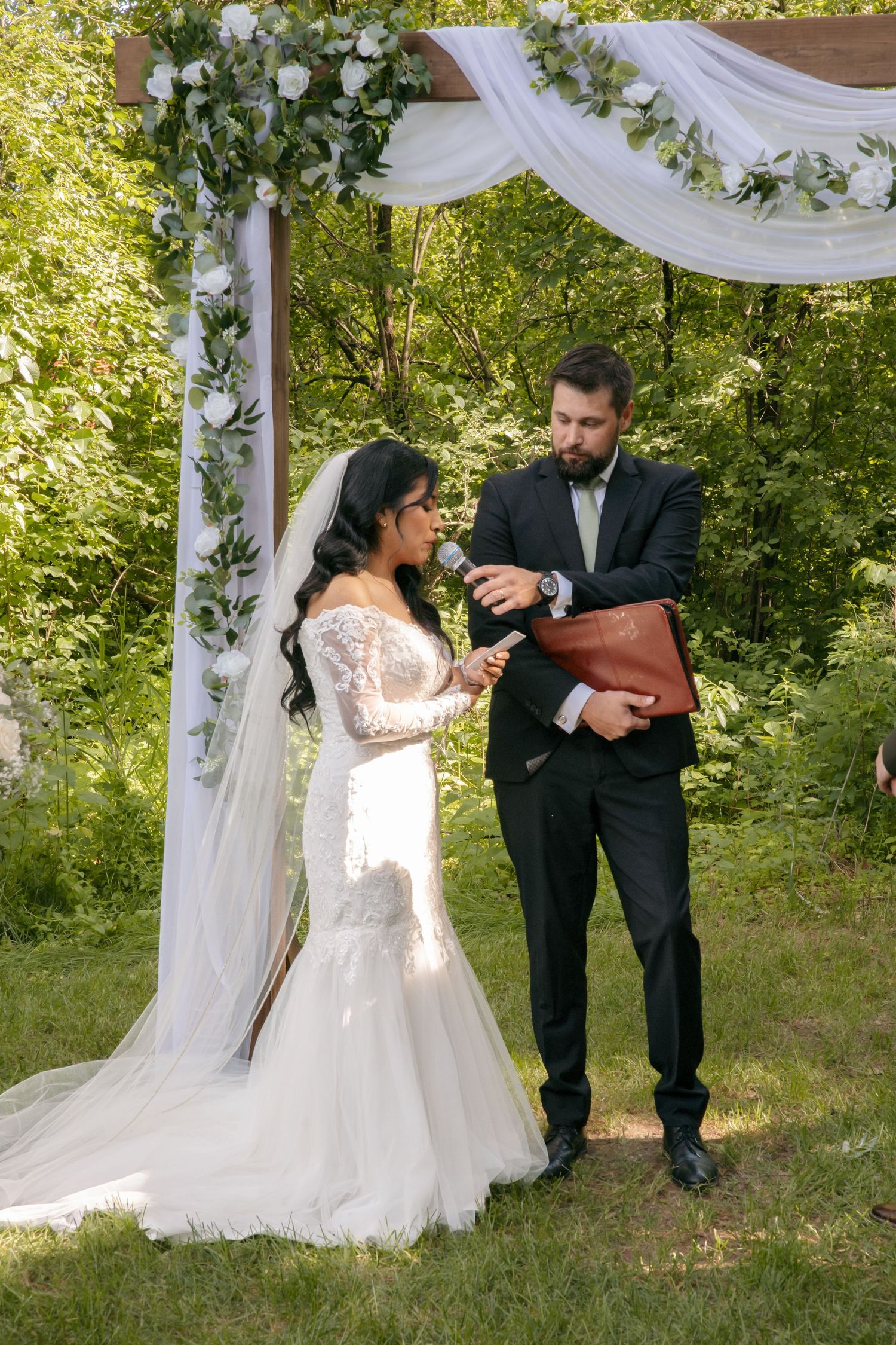 Groom in black tuxedo reads vows with bride in lace mermaid gown under floral arch during outdoor wedding ceremony.