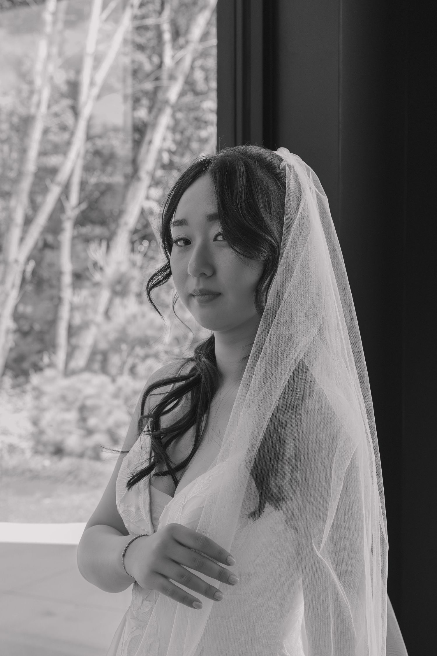 Elegant black and white portrait of a bride in a veil gazing thoughtfully beside a large window.