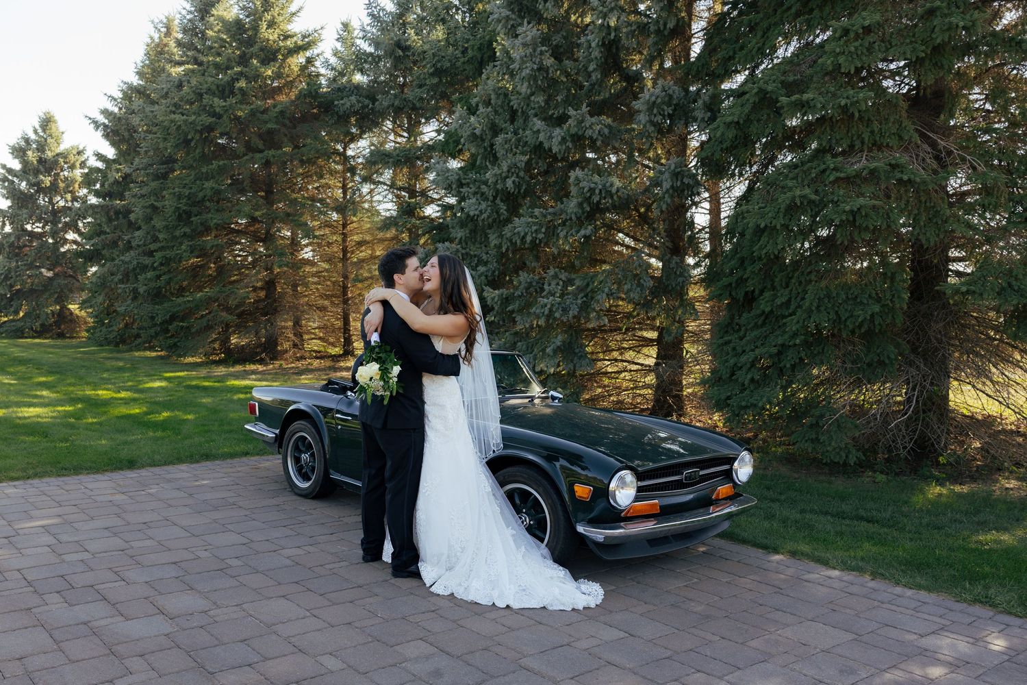 Bride and groom kissing beside a classic black convertible car surrounded by tall pine trees on their wedding day.