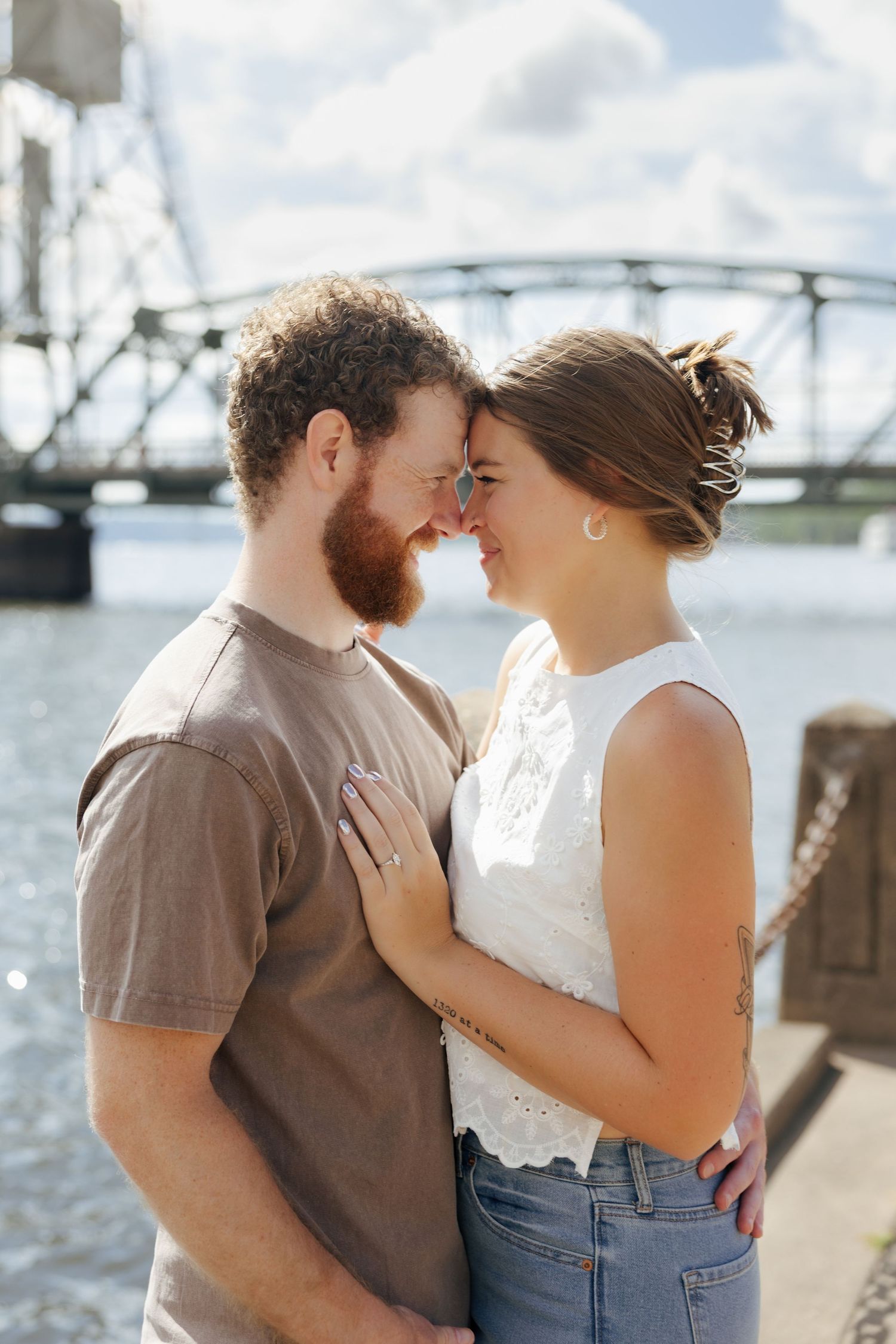 A couple shares a romantic moment by a river with a bridge backdrop, touching foreheads and embracing tenderly.