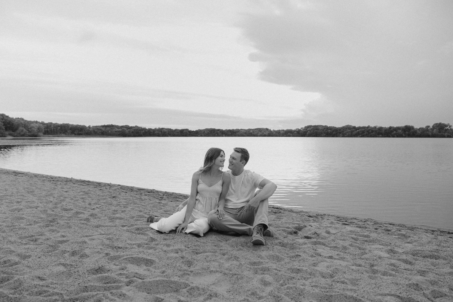 A romantic couple sits close together on a sandy beach by a calm lake in black and white.