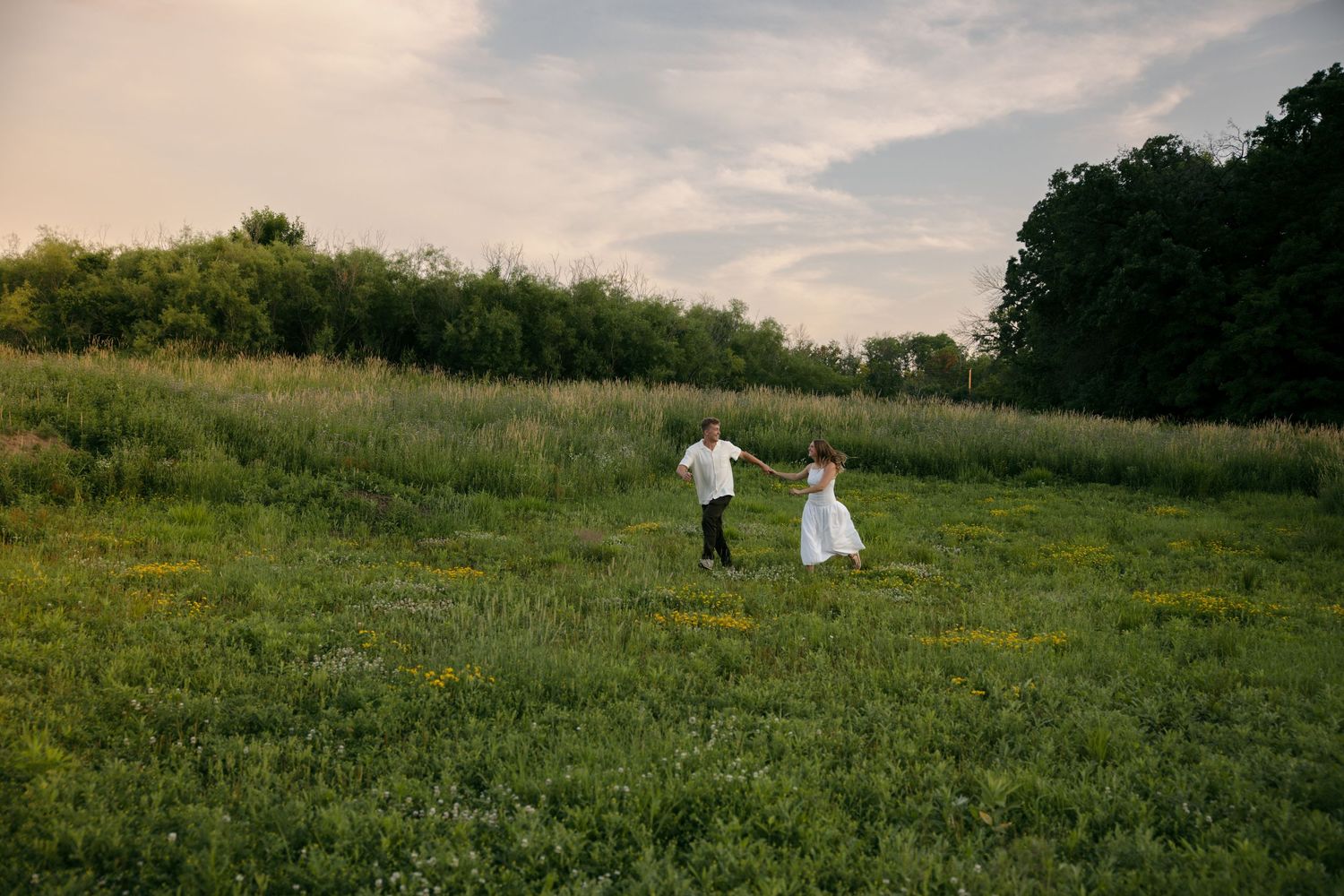Couple holding hands and running playfully through a vast green meadow at sunset.
