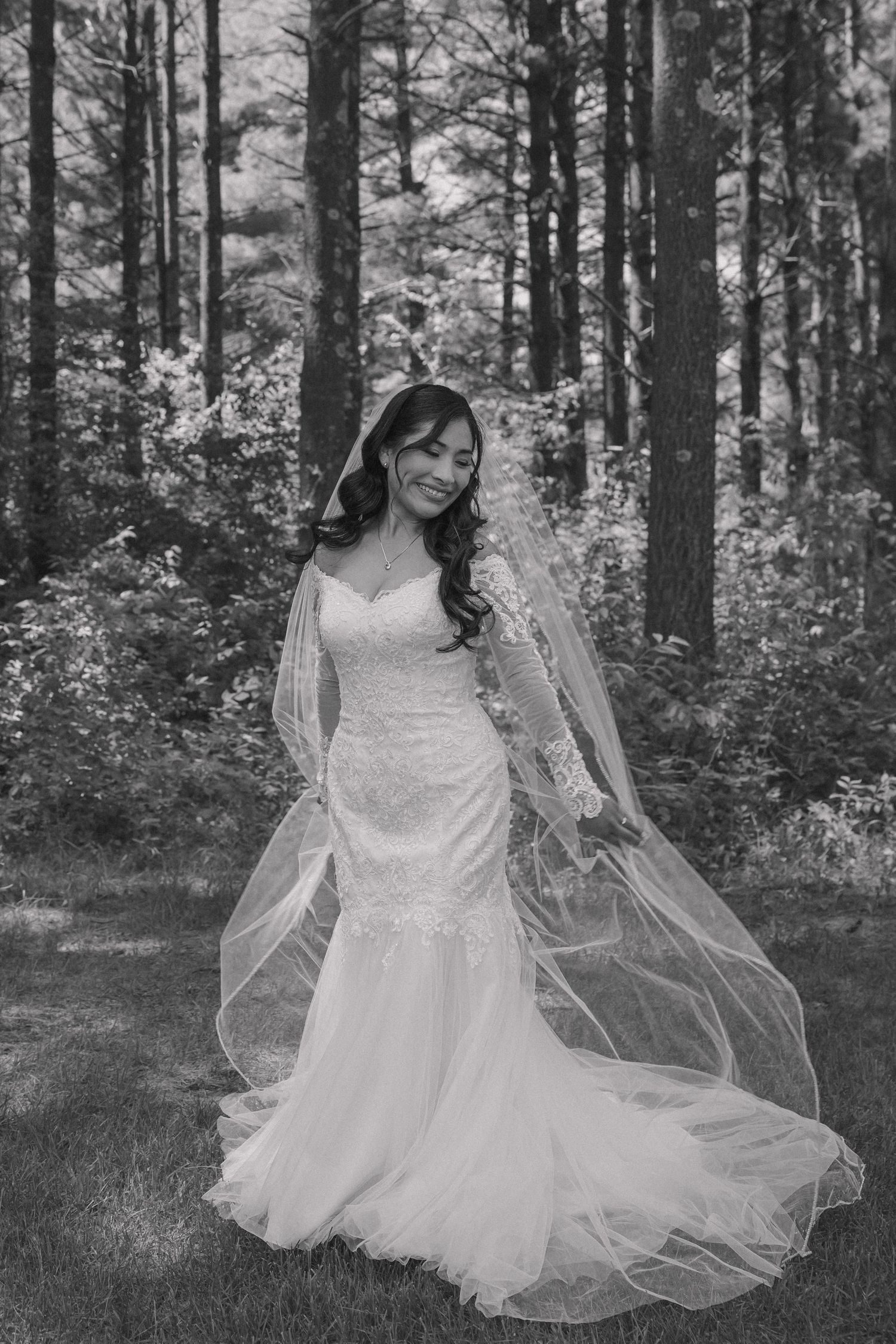 Black and white photo of smiling bride in fitted lace gown with flowing veil in forest.