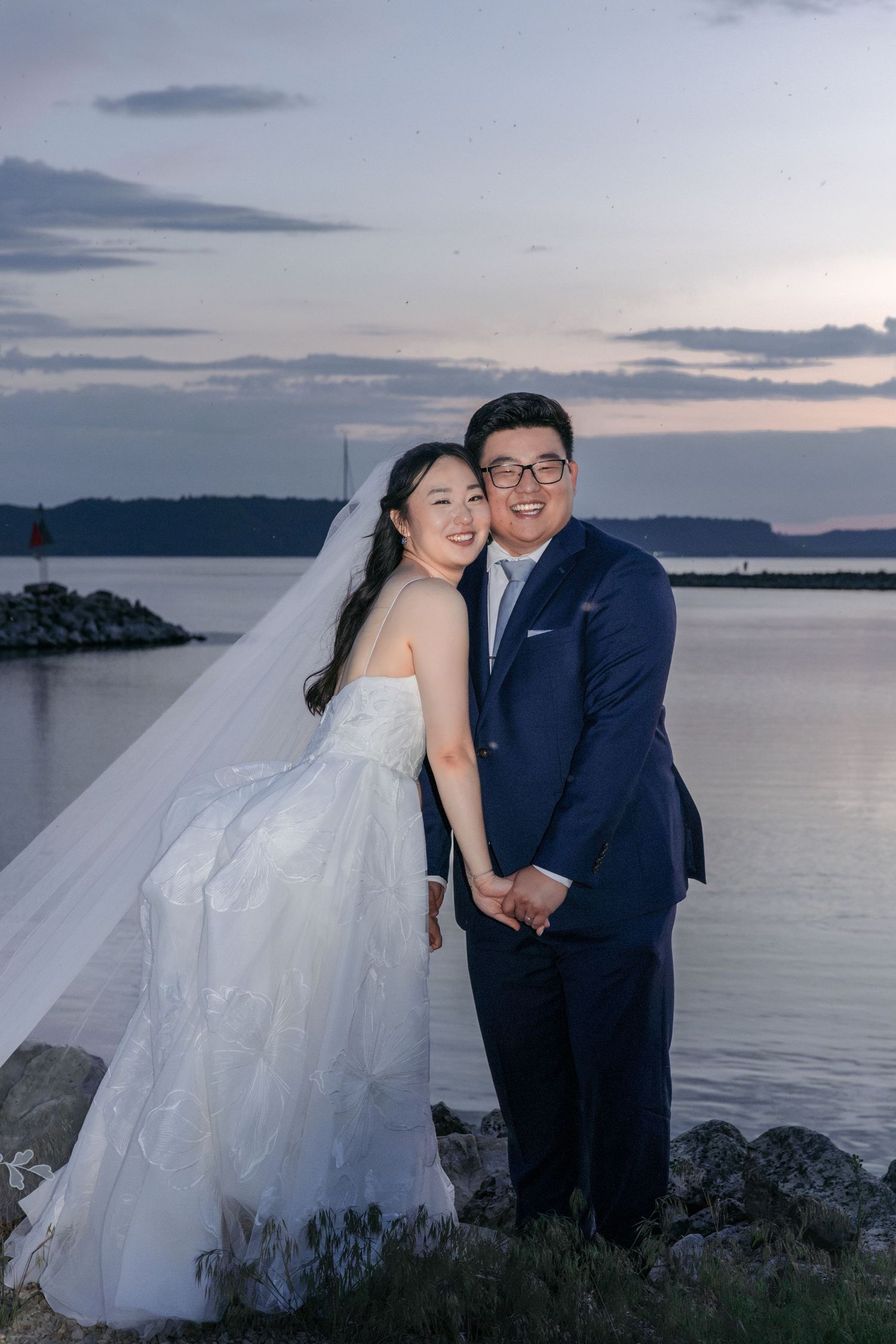 Smiling bride and groom pose together by a serene lake at dusk during their wedding celebration.