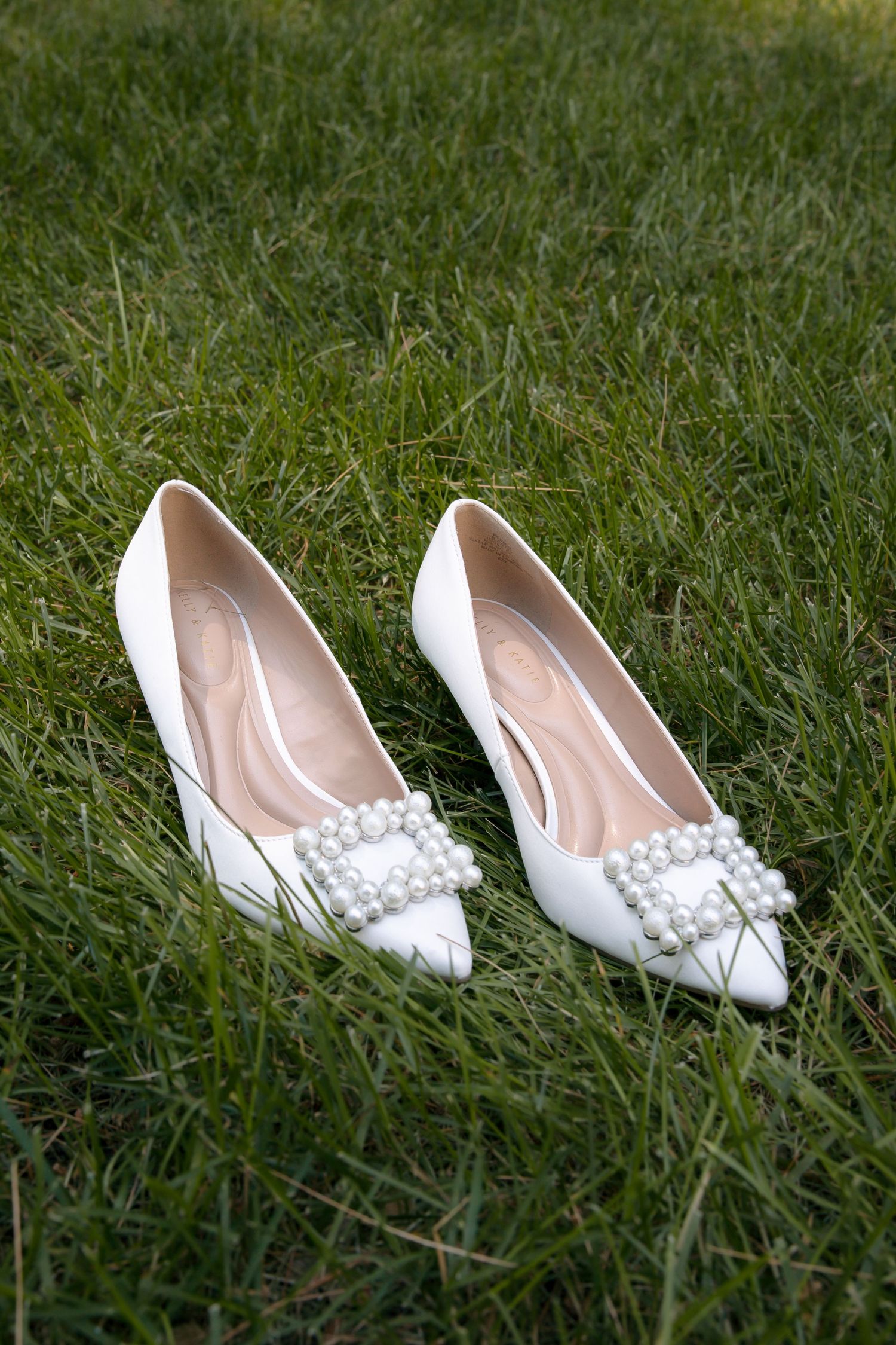 White bridal heels with pearl buckle embellishment resting on green grass, perfect wedding shoes.