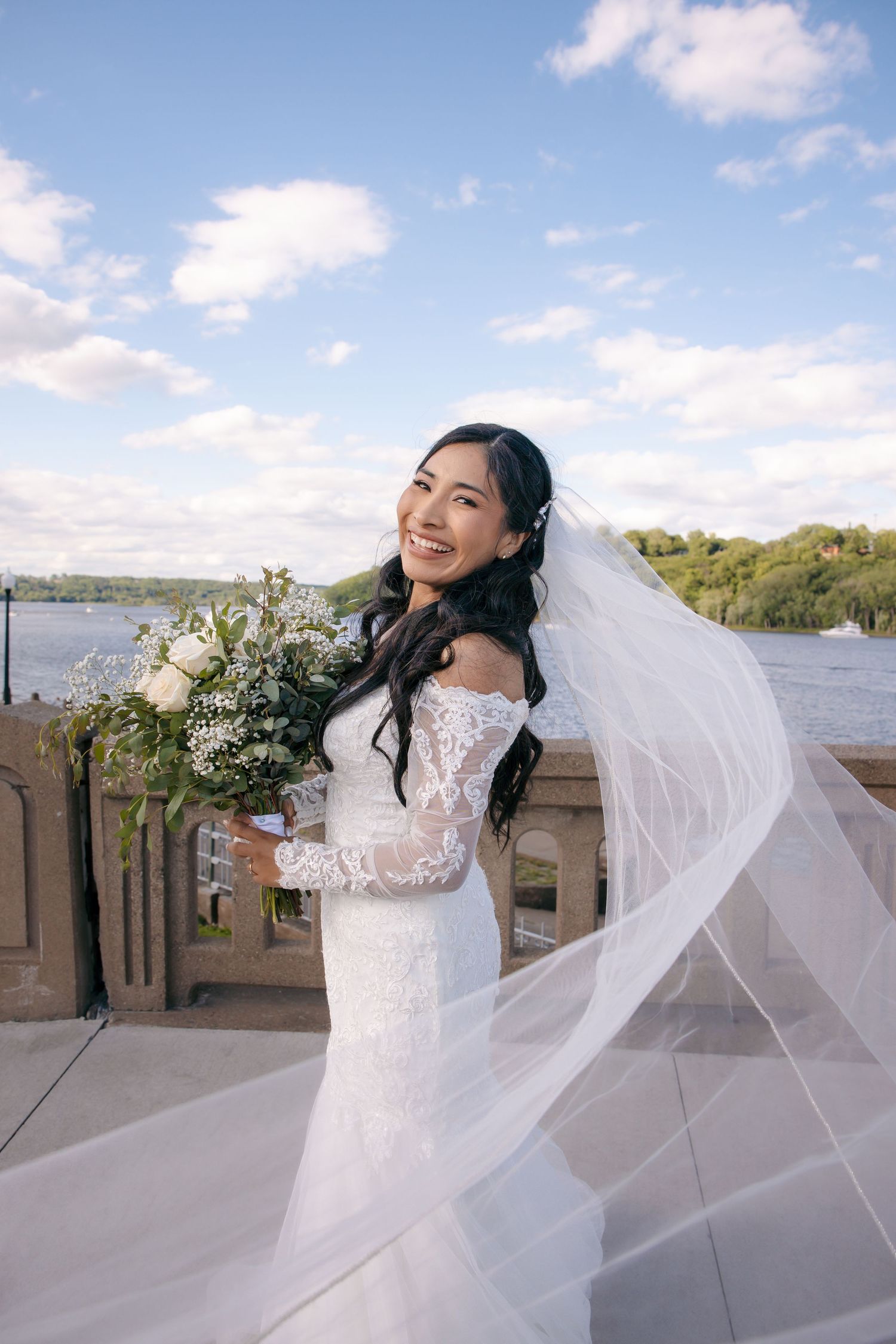 Radiant bride smiles outdoors with veil blowing in wind, holding white bouquet with a lake and blue sky behind her.