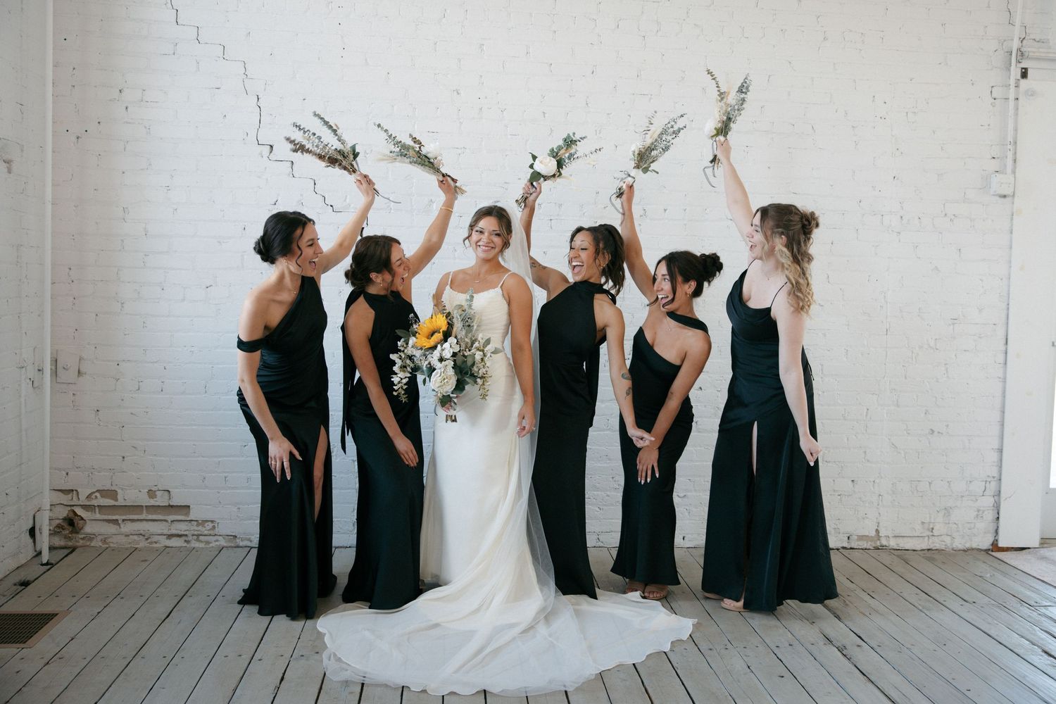 Bride in white gown surrounded by bridesmaids in black dresses raising floral bouquets in bright white brick venue.