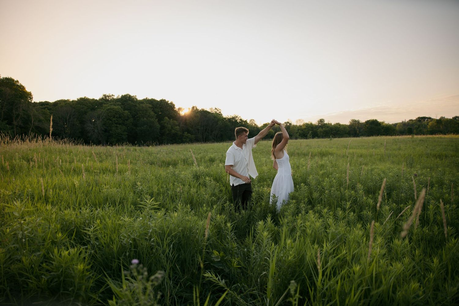 Couple dancing in a lush green field at golden hour sunset with trees in the background.